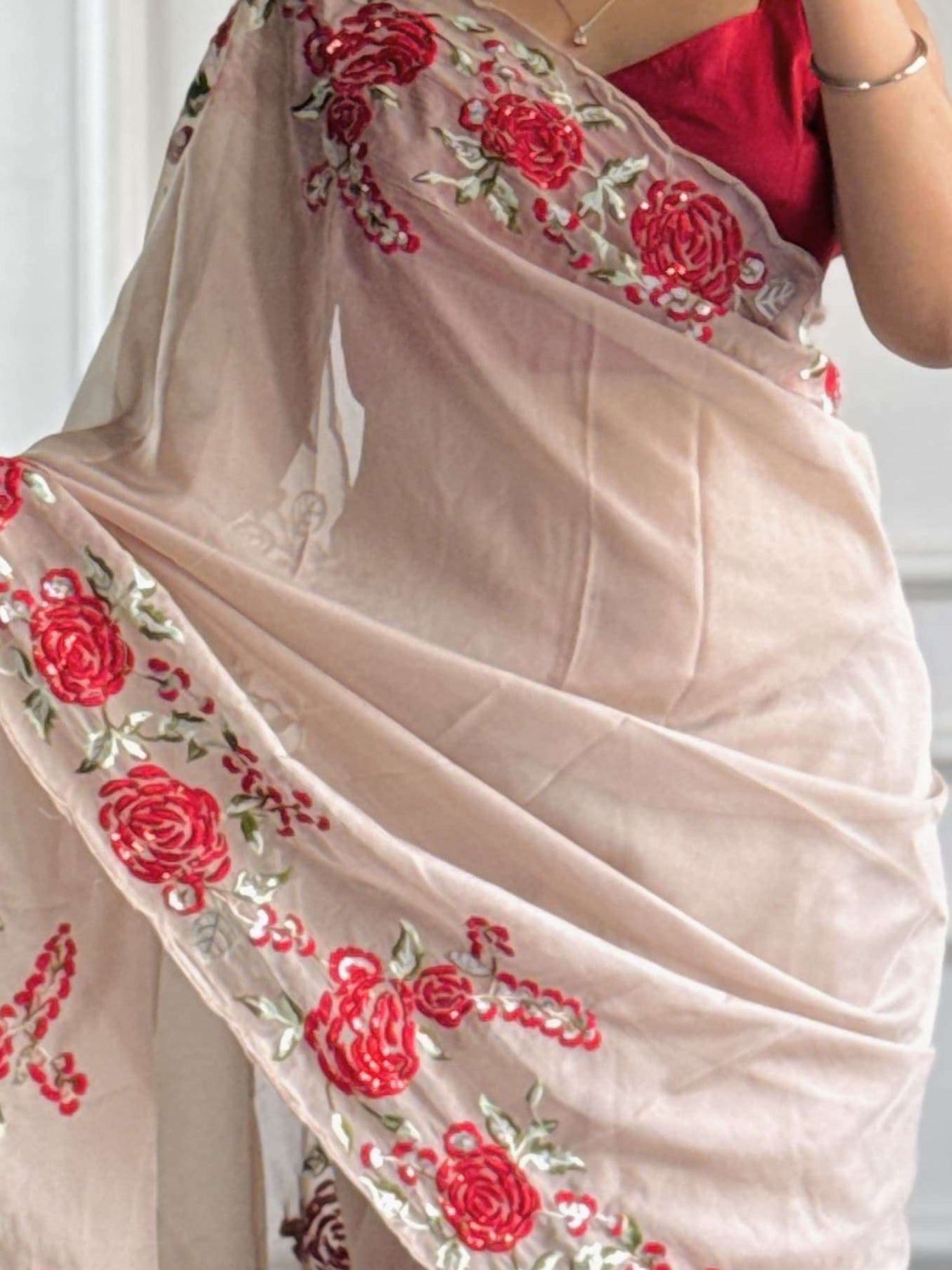 Woman wearing a beige saree with red floral patterns in front of a white paneled wall.
