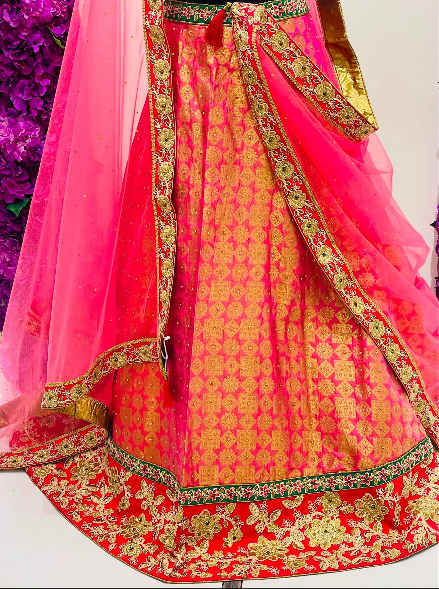 A pink and red lehenga Skirt with intricate gold and green zari embroidery, featuring a matching sheer pink dupatta draped over the side.