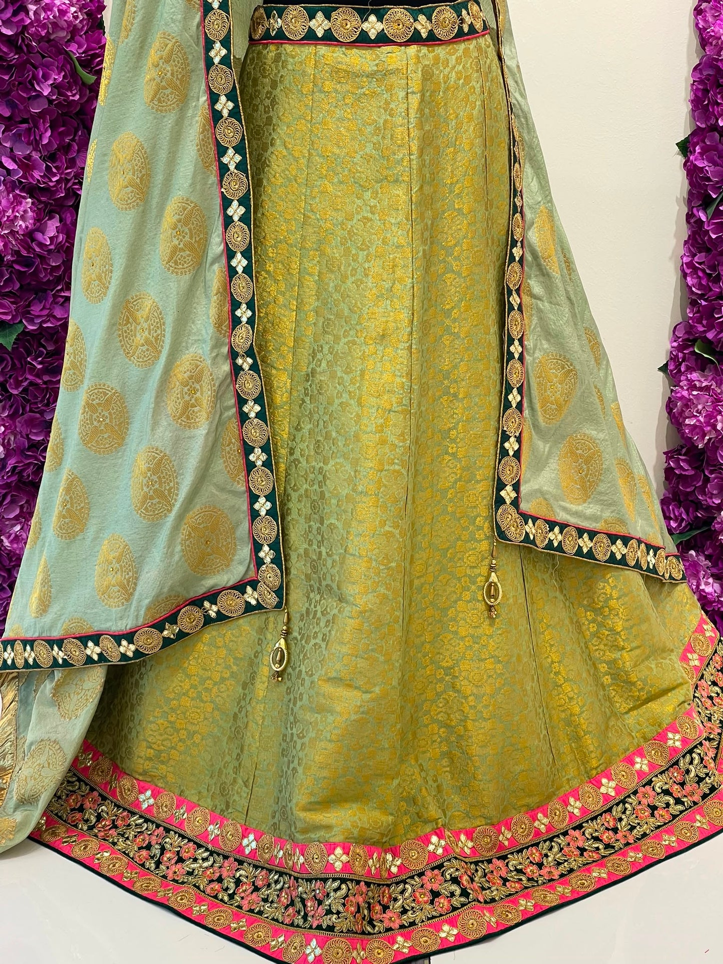 A traditional Indian ethnic outfit featuring a light olive green flared skirt with intricate gold and pink borders, paired with a light blue-green dupatta decorated with gold motifs and black and gold patterned borders.