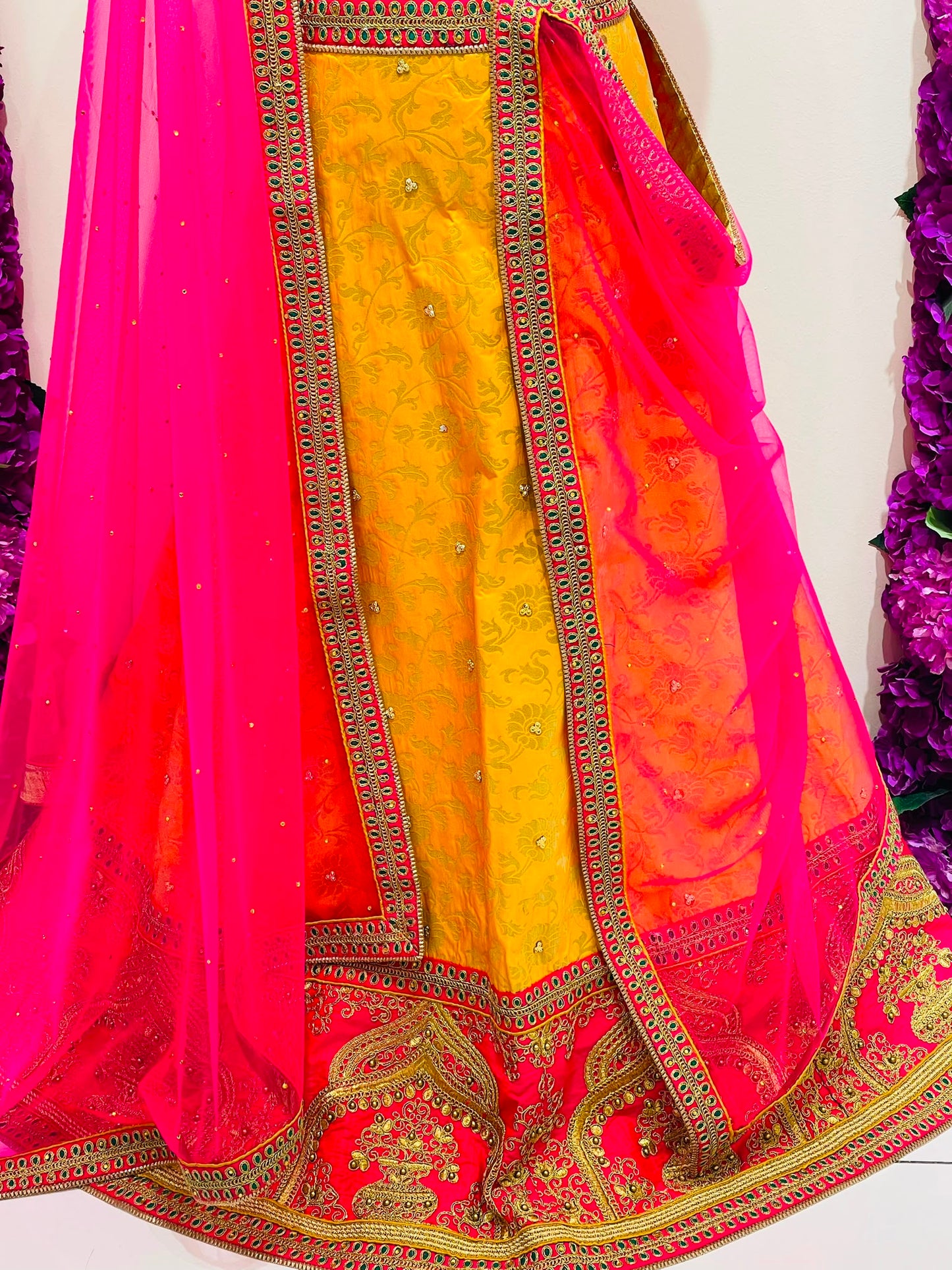 A close-up of a traditional Indian outfit featuring a bright yellow embroidered kurta with a pink and orange gradient dupatta and a vibrant pink border, decorated with gold zari embroidery.