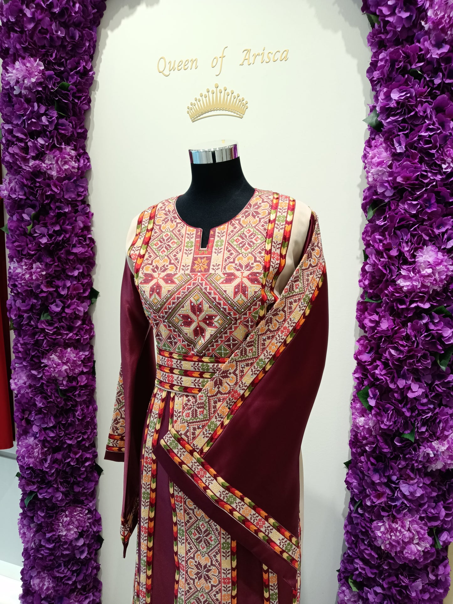 rich maroon satin-finish fabric, this dress features intricate multicolor embroidery inspired by traditional Palestinian patterns, creating a timeless and sophisticated look.
