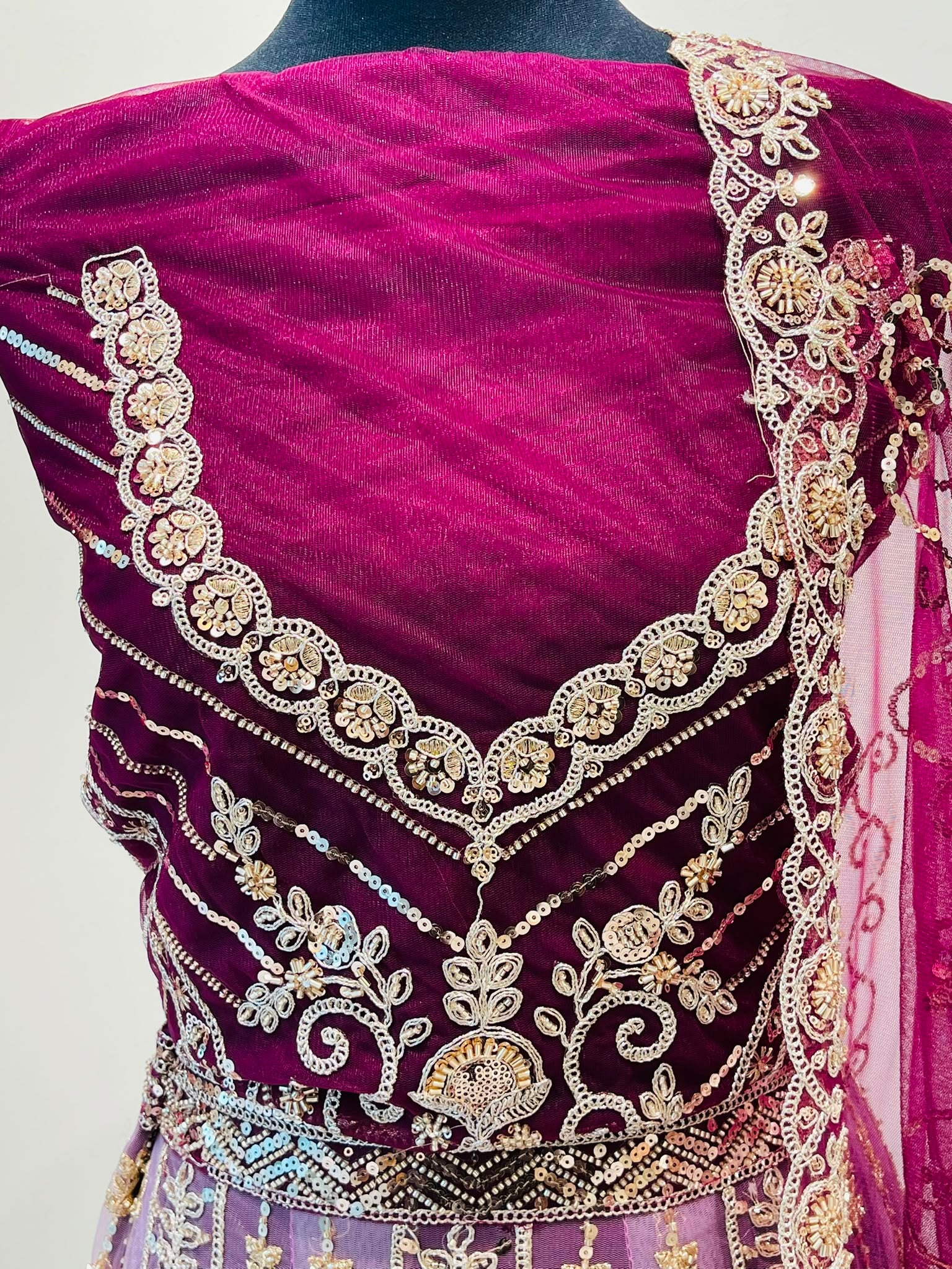 A close-up of a magenta velvet and mesh lehenga garment with extensive gold and silver embroidery, beadwork, and floral motifs.