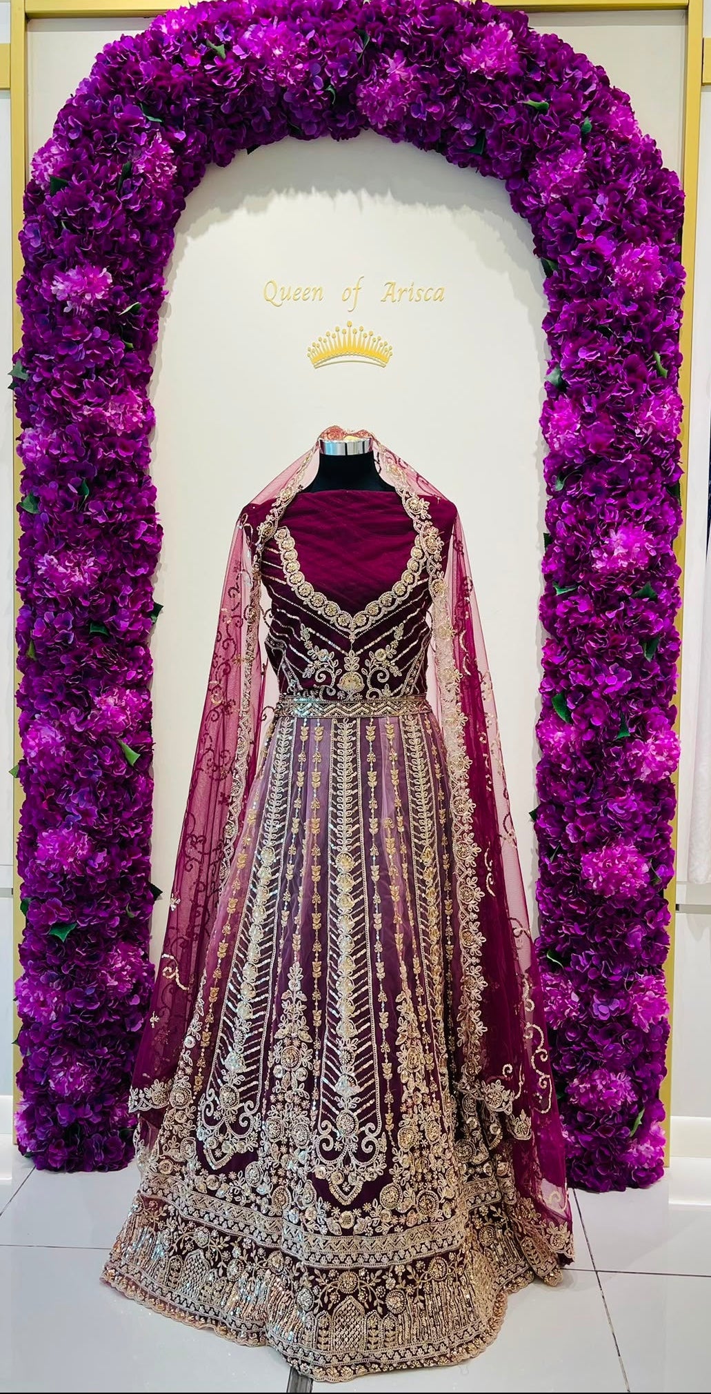 An ornate traditional Indian lehenga choli dress displayed on a mannequin inside Arisca boutique. The dress is deep purple with heavy gold and silver embroidery, paired with a sheer purple dupatta.