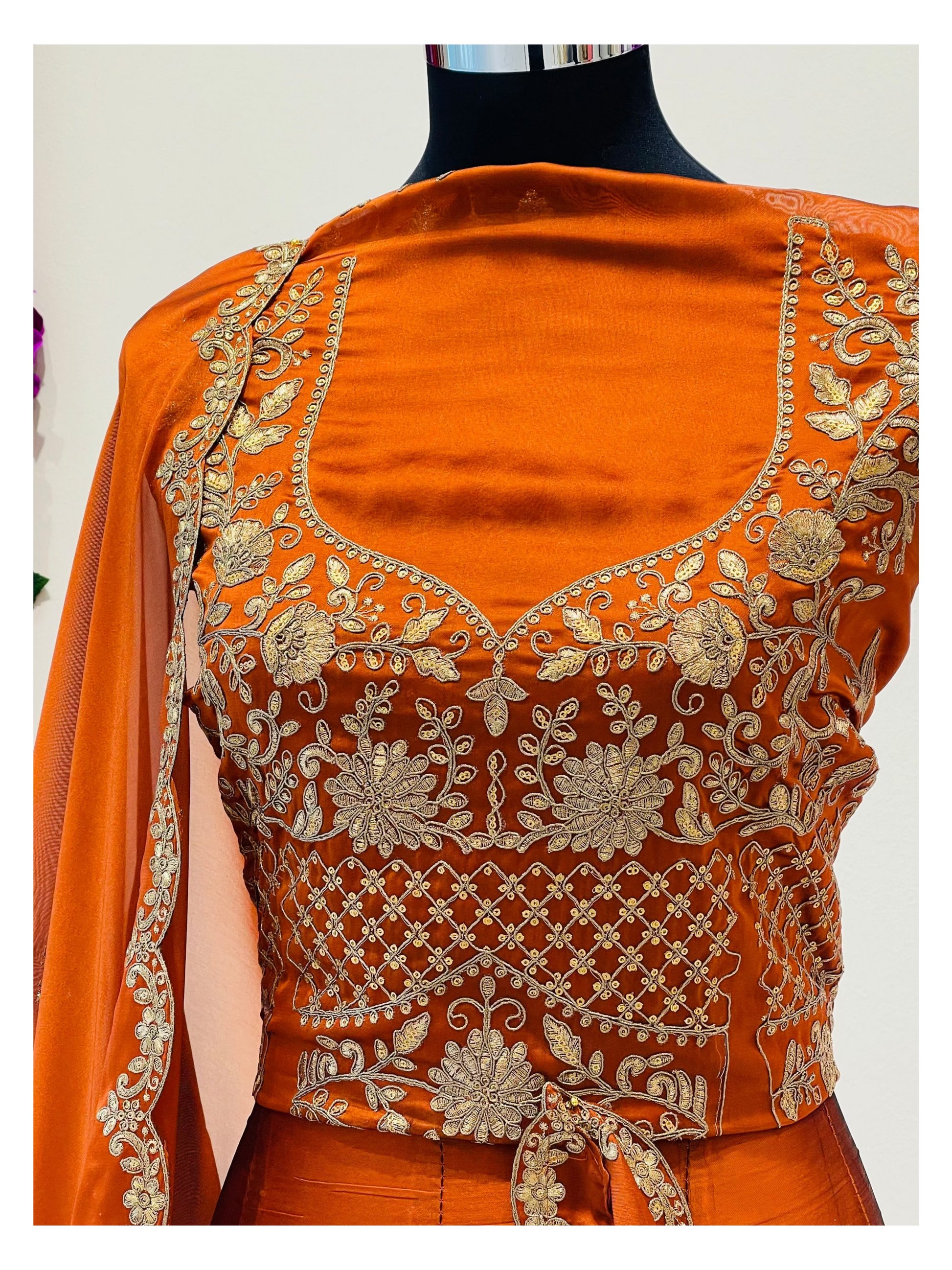 An orange embroidered traditional blouse  , featuring intricate gold floral and geometric embroidery, paired with a matching orange dupatta draped over one shoulder.