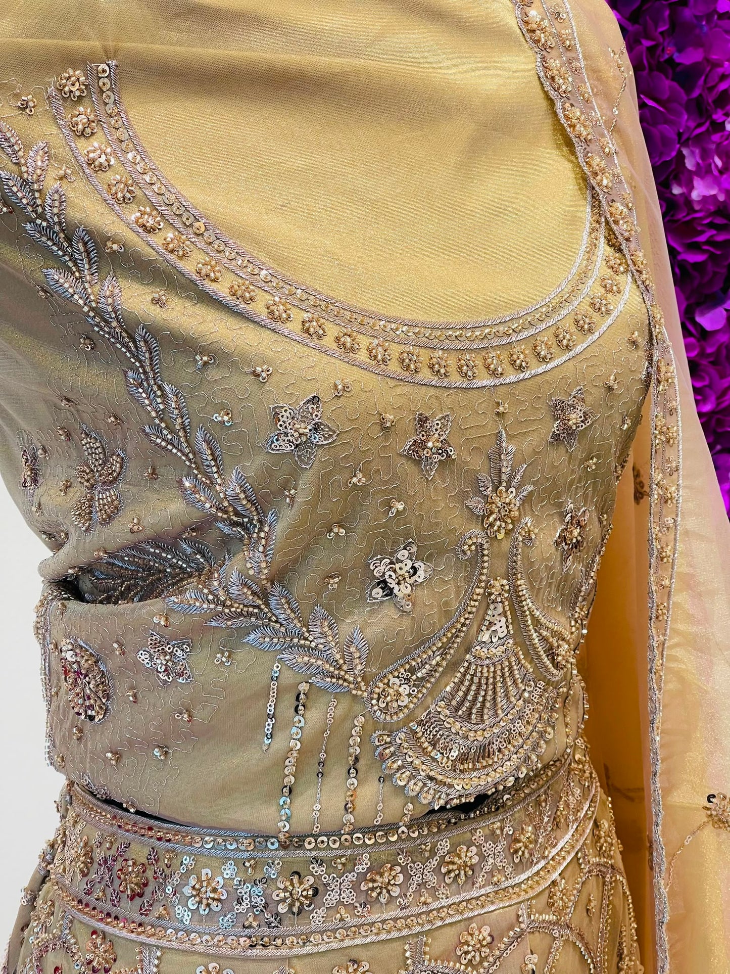 Close-up of a beige or champagne-colored embroidered traditional lehenga with intricate silver and gold metallic beadwork, sequins, and floral motifs. A matching sheer dupatta is draped over the shoulder