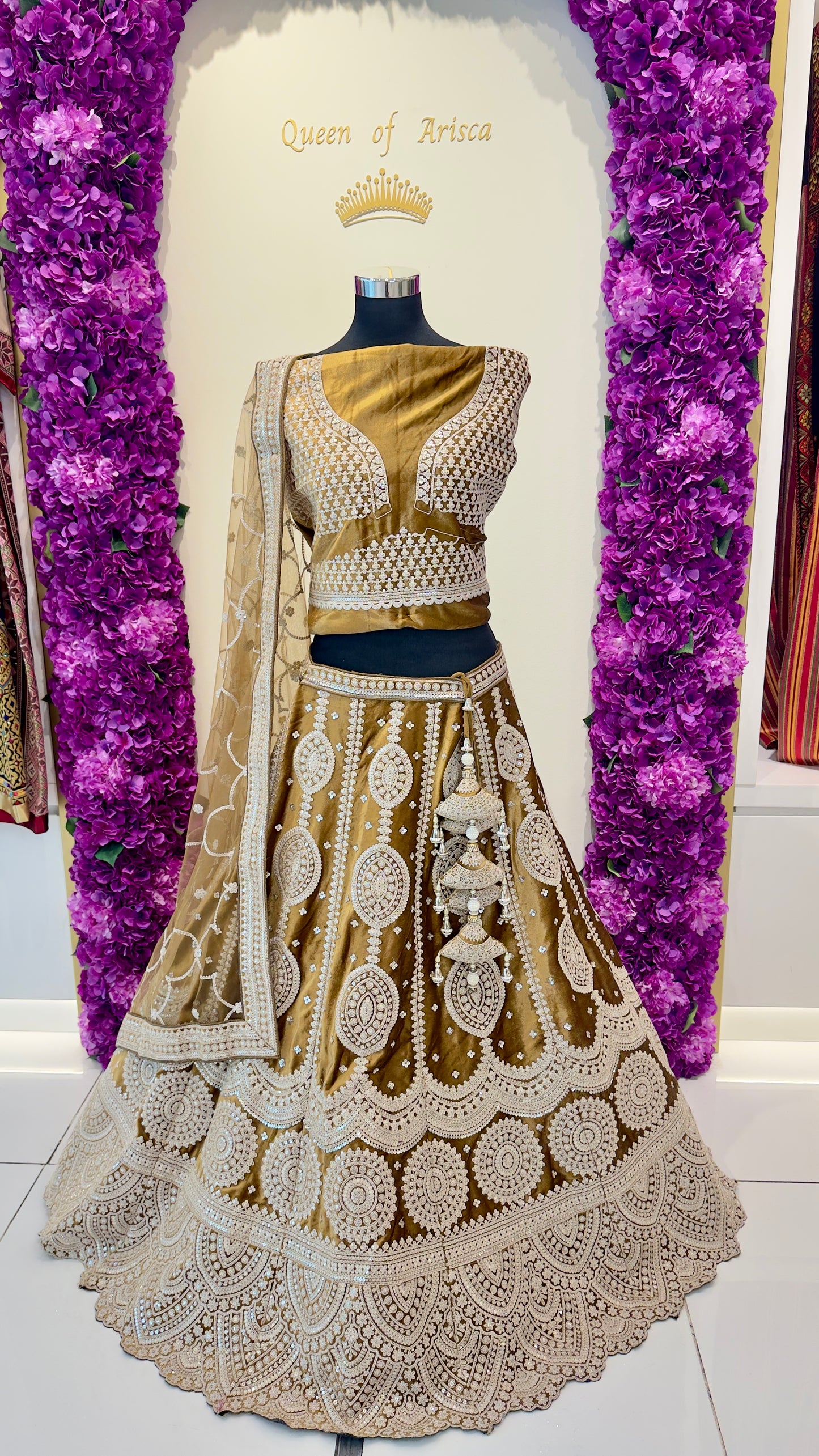 Traditional embroidered lehenga with gold and white design on a mannequin against a purple floral backdrop.