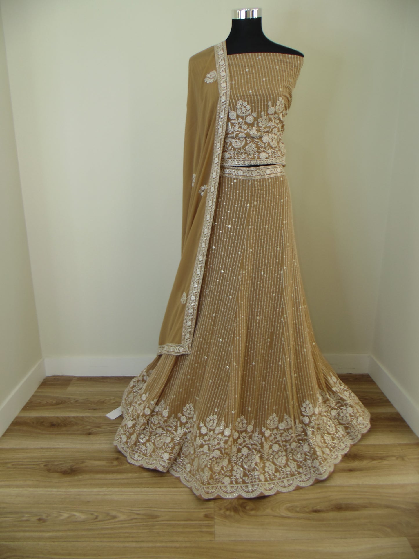 Beige evening Lehenga with embroidery details on a wooden stand against a white wall.