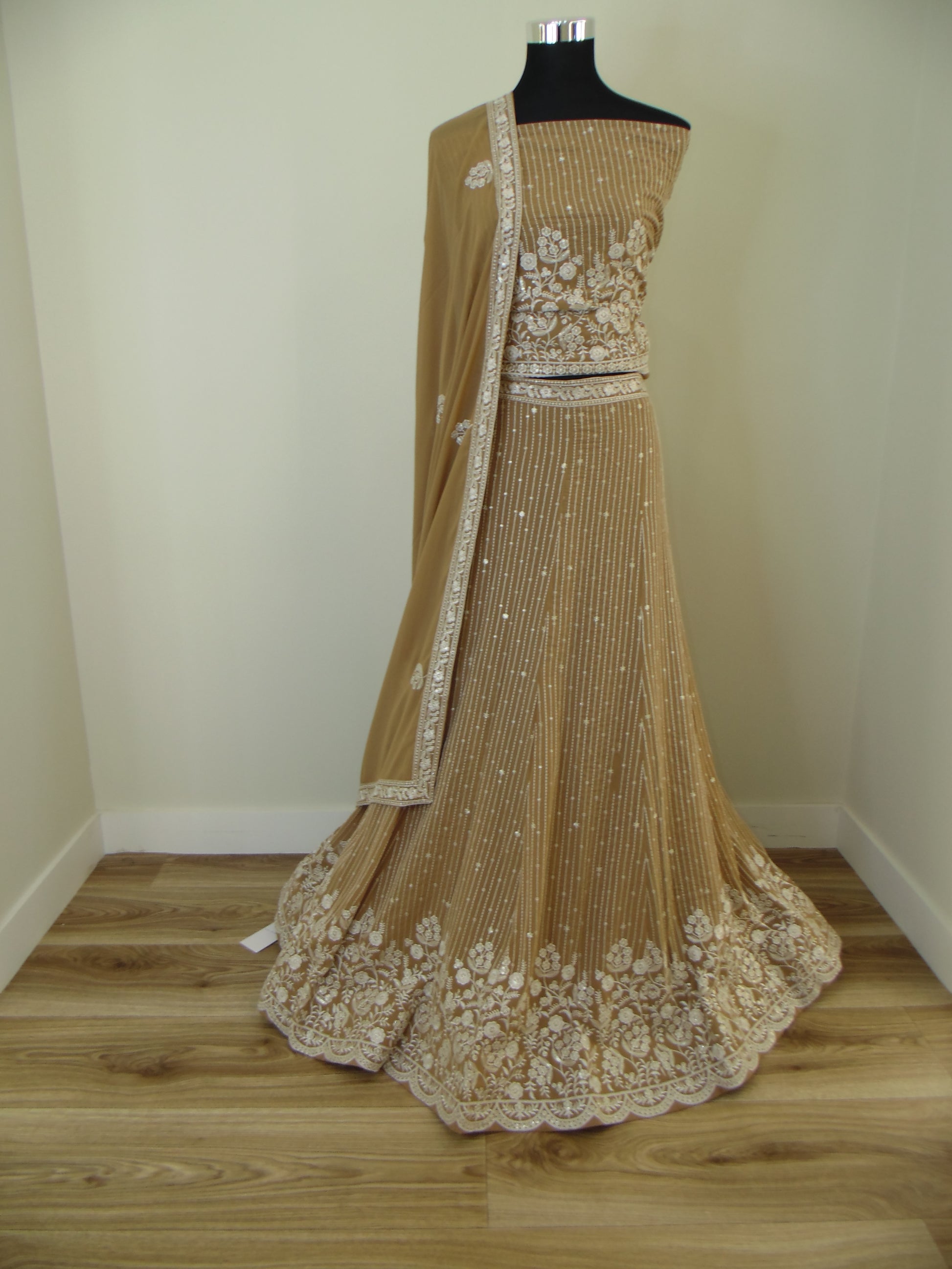 Beige evening Lehenga with embroidery details on a wooden stand against a white wall.