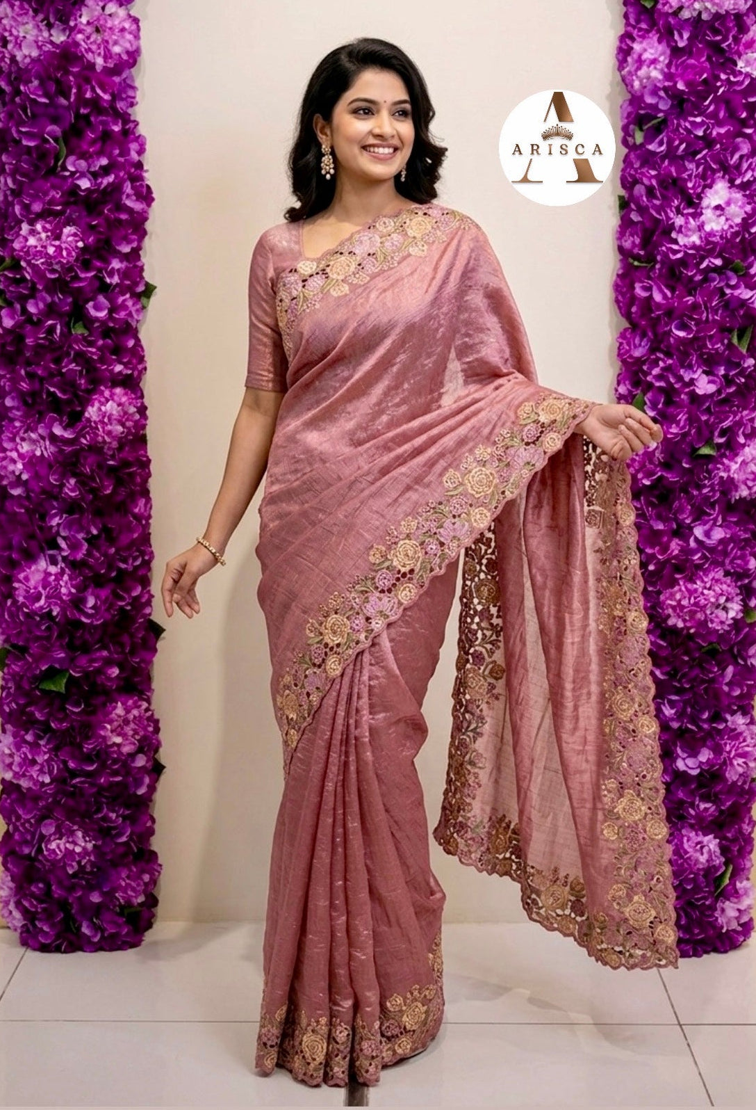 Woman in a pink saree with gold embroidery standing in front of purple floral decorations.