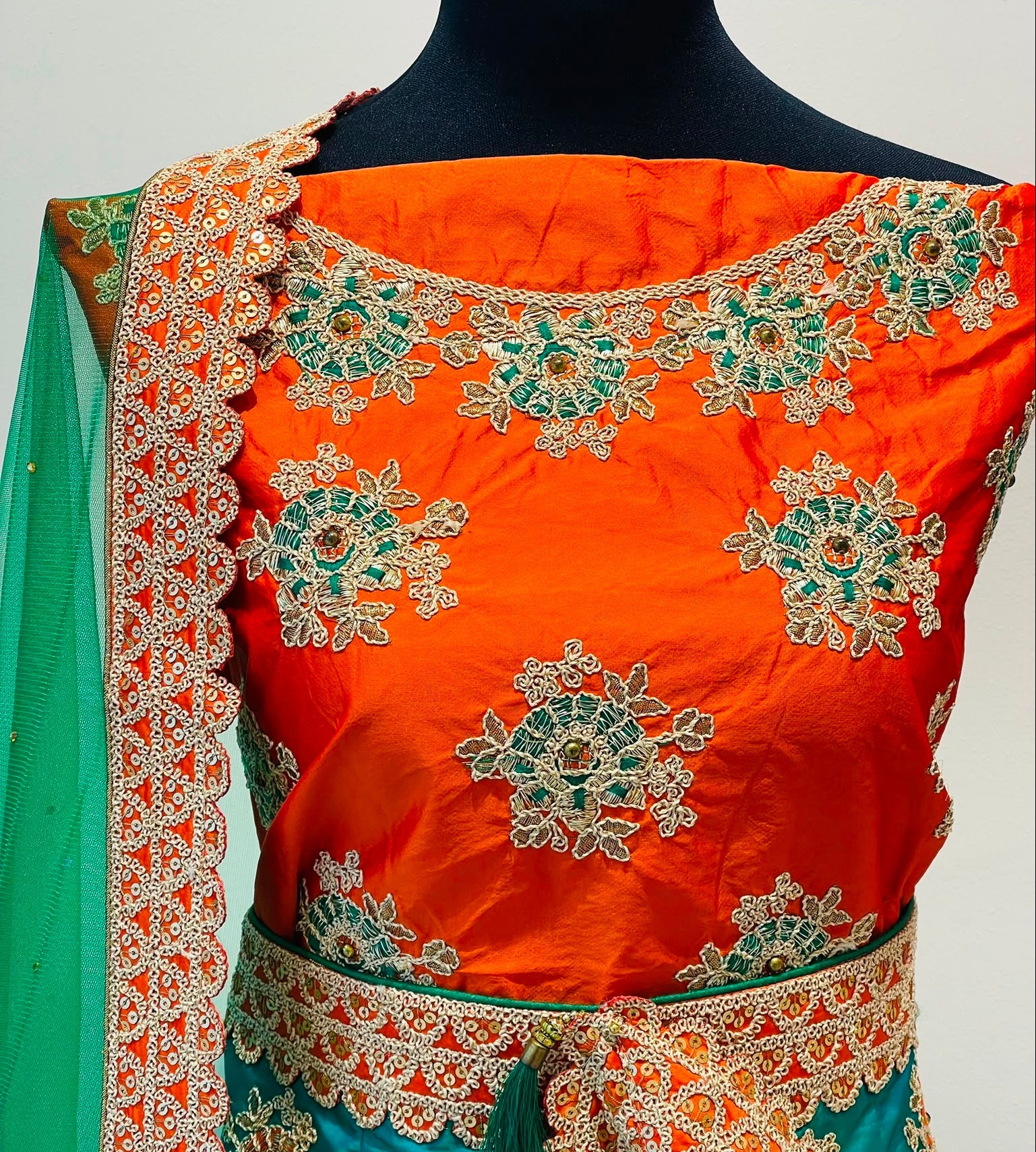 An orange and teal embroidered traditional Indian outfit . The blouse is bright orange with intricate green and gold floral embroidery, paired with a teal skirt and a matching green dupatta draped over the shoulder.
