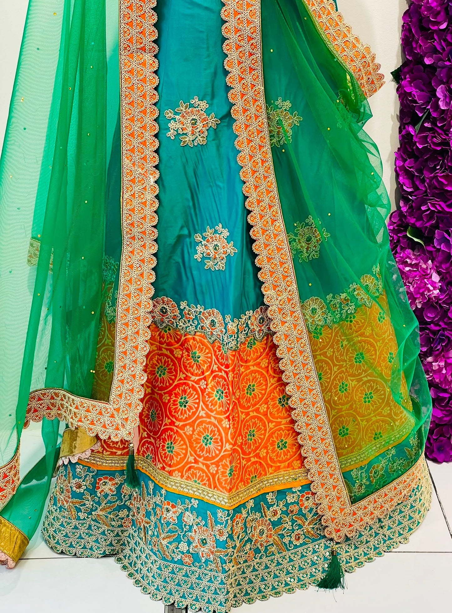 A close-up of a traditional Indian lehenga , featuring a teal blue skirt with a wide orange and yellow embroidered border, a matching teal dupatta, and intricate gold and silver embroidery.