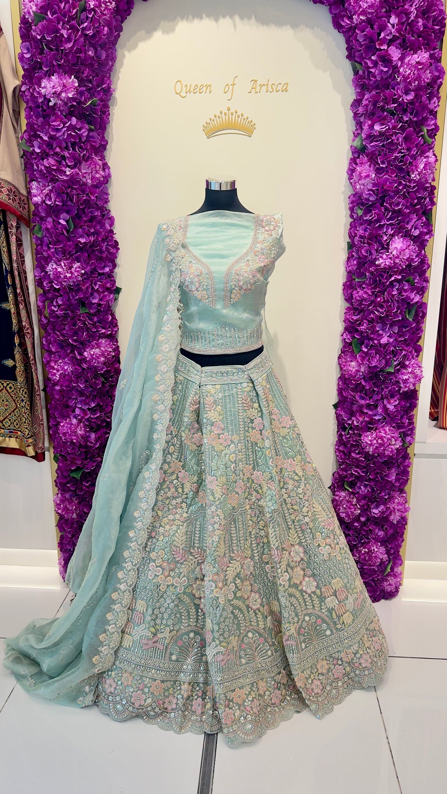 Light green embroidered lehenga with matching blouse on a mannequin against a purple floral backdrop.