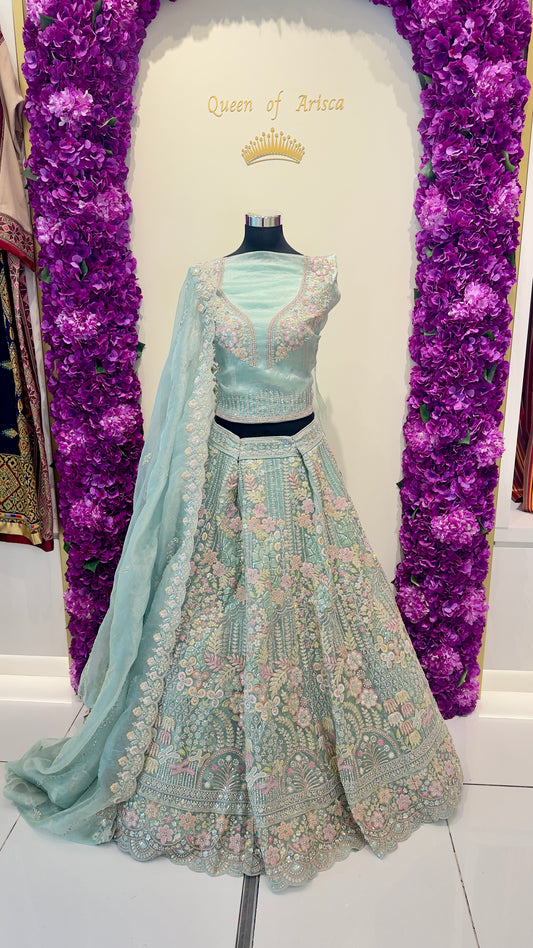 Light green embroidered lehenga with matching blouse on a mannequin against a purple floral backdrop.
