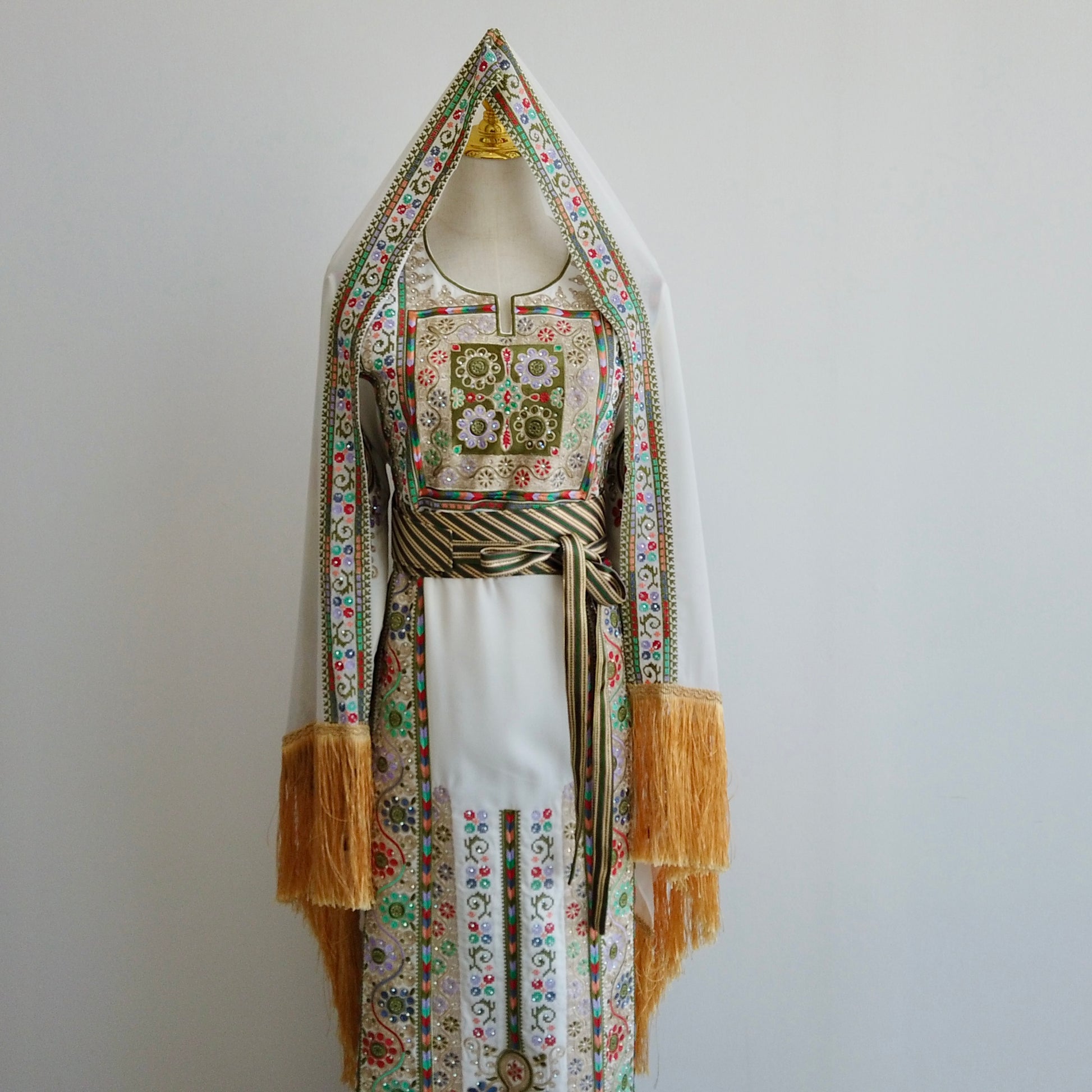 Front-facing view of embroidered Palestinian thobe with magenta, green, and white motifs. displayed on mannequin with striped waist belt.