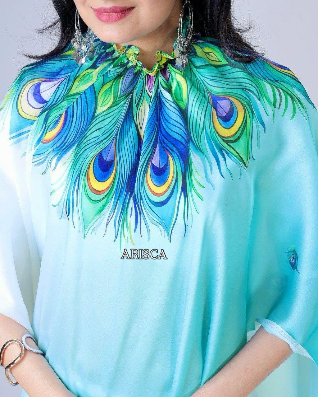 Person wearing a turquoise blouse with peacock feather design, brand 'ARISCA' visible.