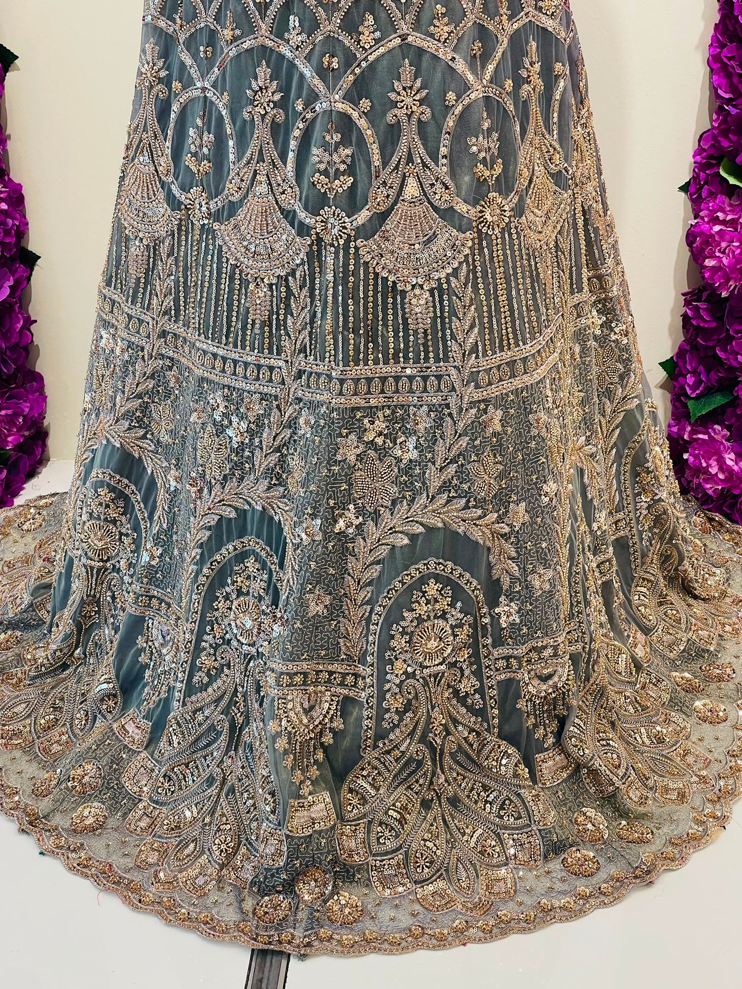 An ornate heavily embroidered traditional slehenga in dark teal-blue mesh with intricate gold metallic beadwork, sequins, and mirror work, displayed against a light background with purple floral decorations on the sides.