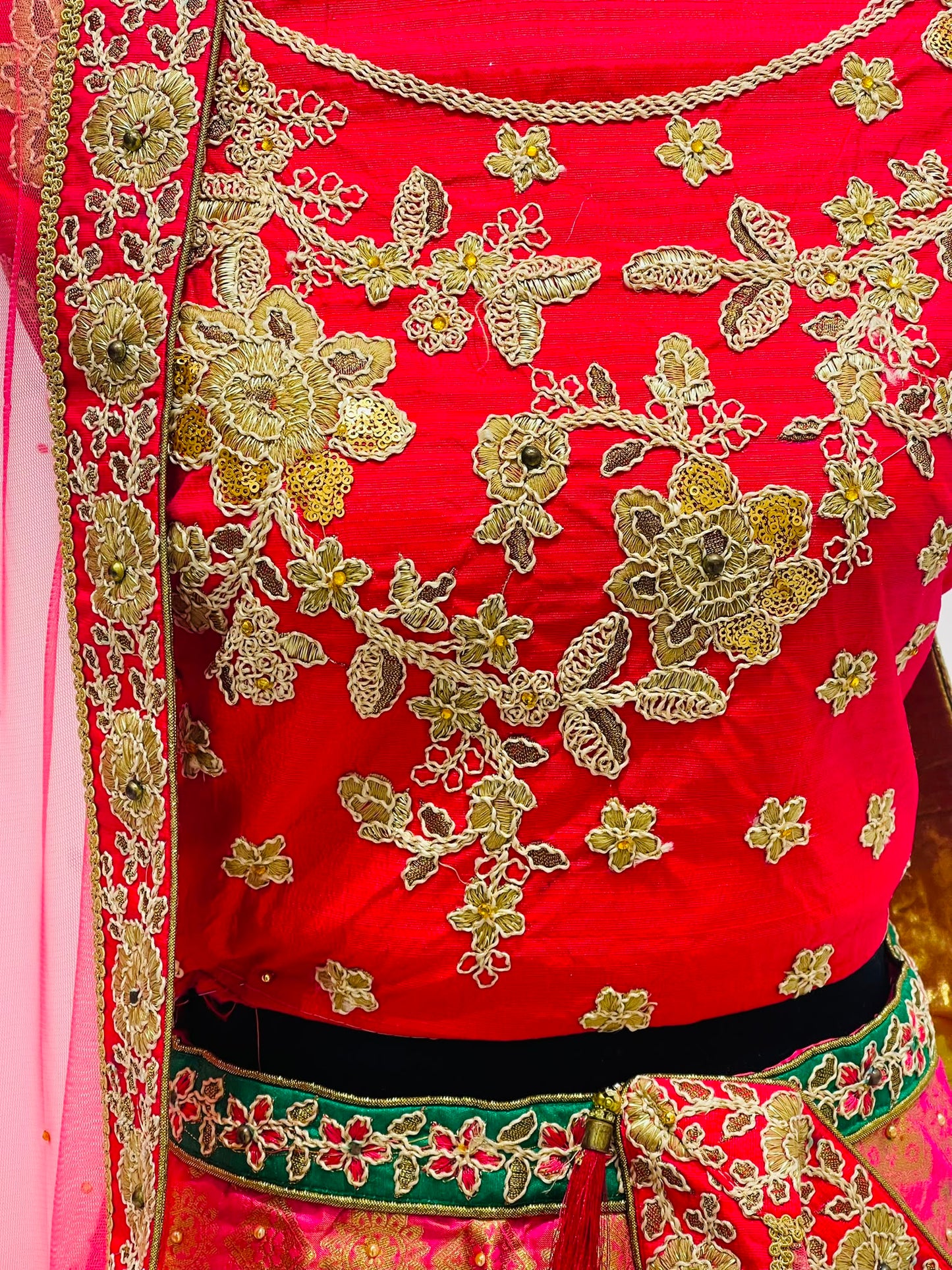 A close-up of a bright red traditional embroidered  blouse of lehenga, featuring intricate gold floral embroidery, and a green and gold border with a matching red dupatta draped on the left.