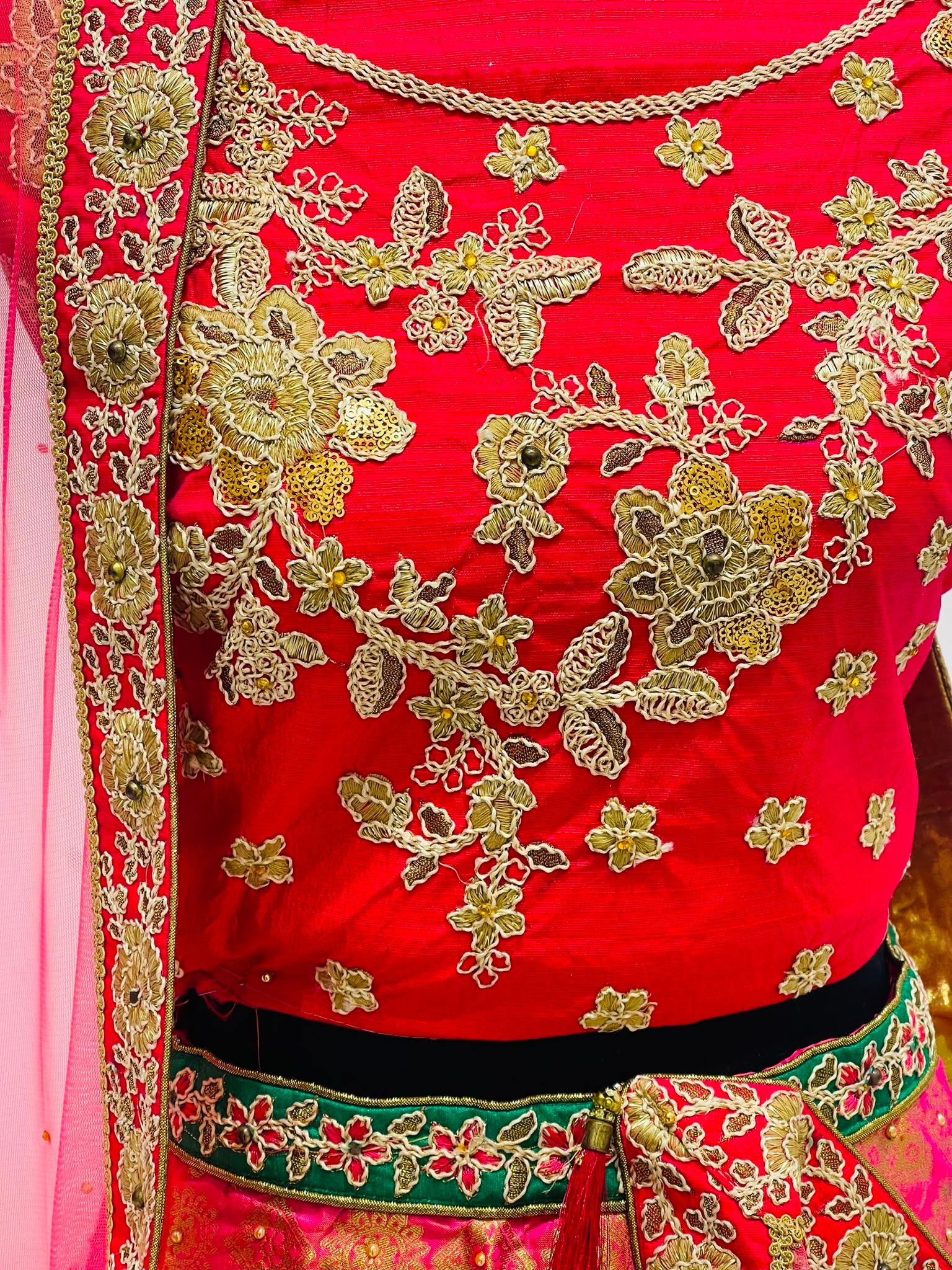 A close-up of a bright red traditional embroidered  blouse of lehenga, featuring intricate gold floral embroidery, and a green and gold border with a matching red dupatta draped on the left.