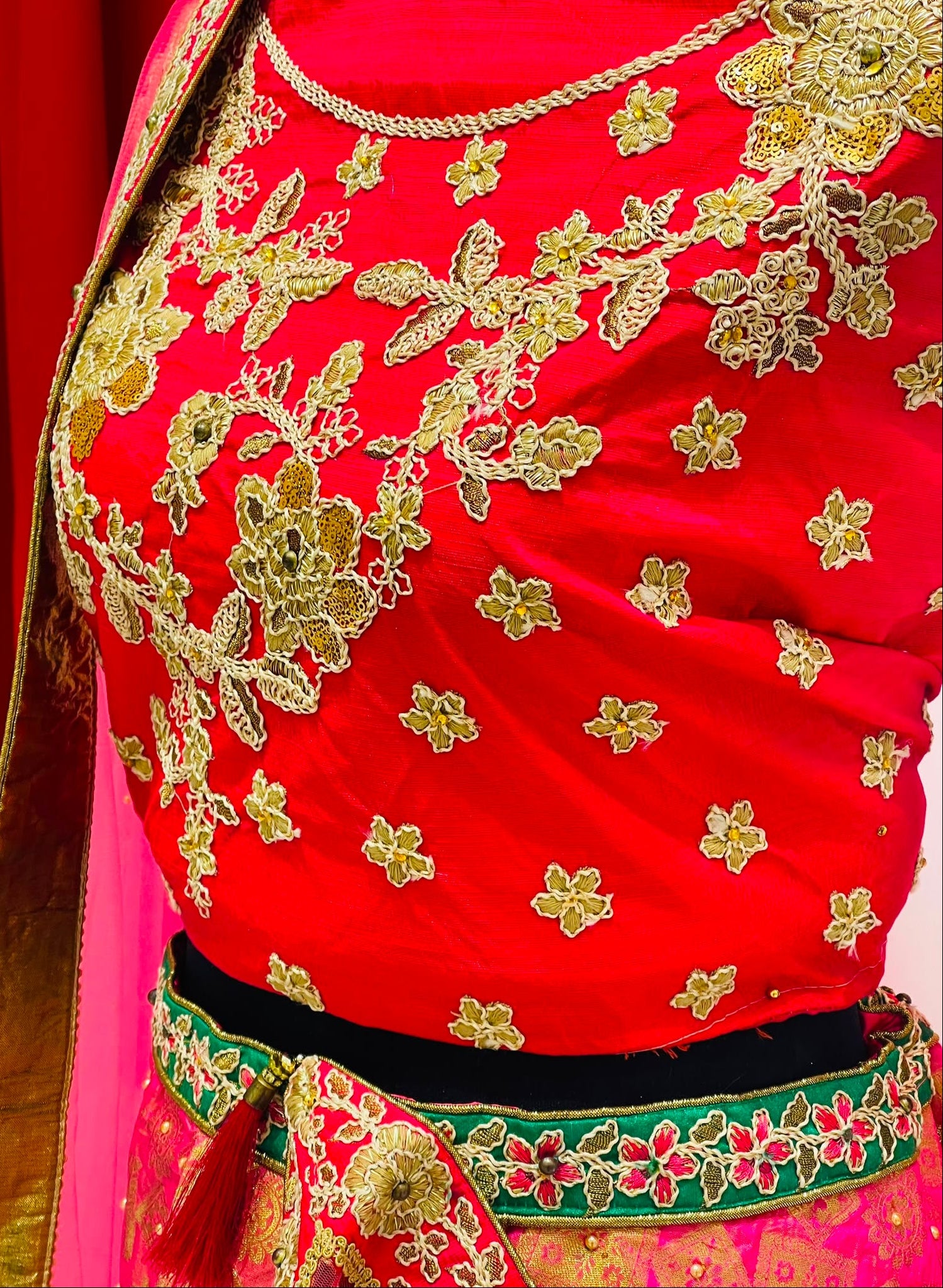 A close-up of a bright red traditional lehenga blouse with extensive gold floral embroidery, and a green and gold embroidered border.