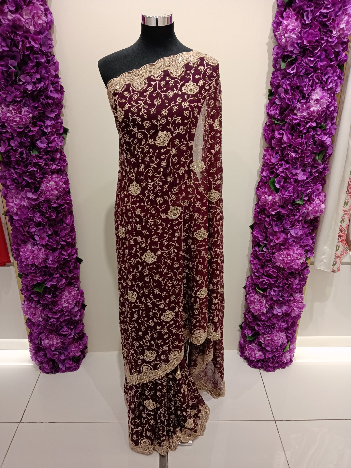 Maroon and gold embroidered saree on a mannequin with purple floral decorations on either side.