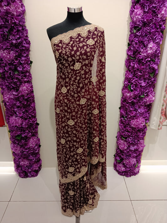 Maroon and gold embroidered saree on a mannequin with purple floral decorations on either side.