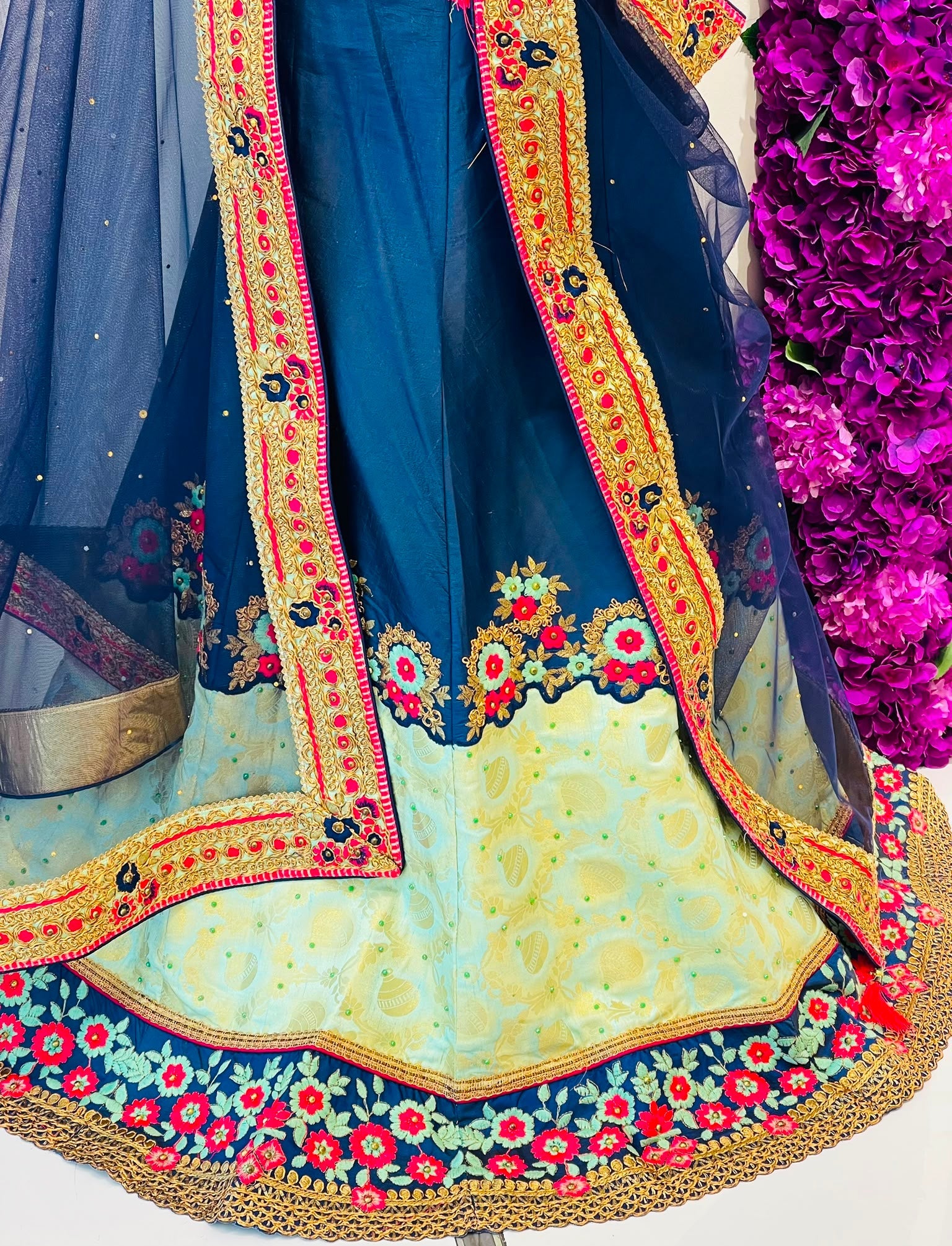 A close-up of a traditional Indian lehenga Skirt featuring a navy blue fabric with a light yellow-green lower panel, heavily decorated with colorful floral embroidery and gold zari borders, alongside a sheer navy blue dupatta.