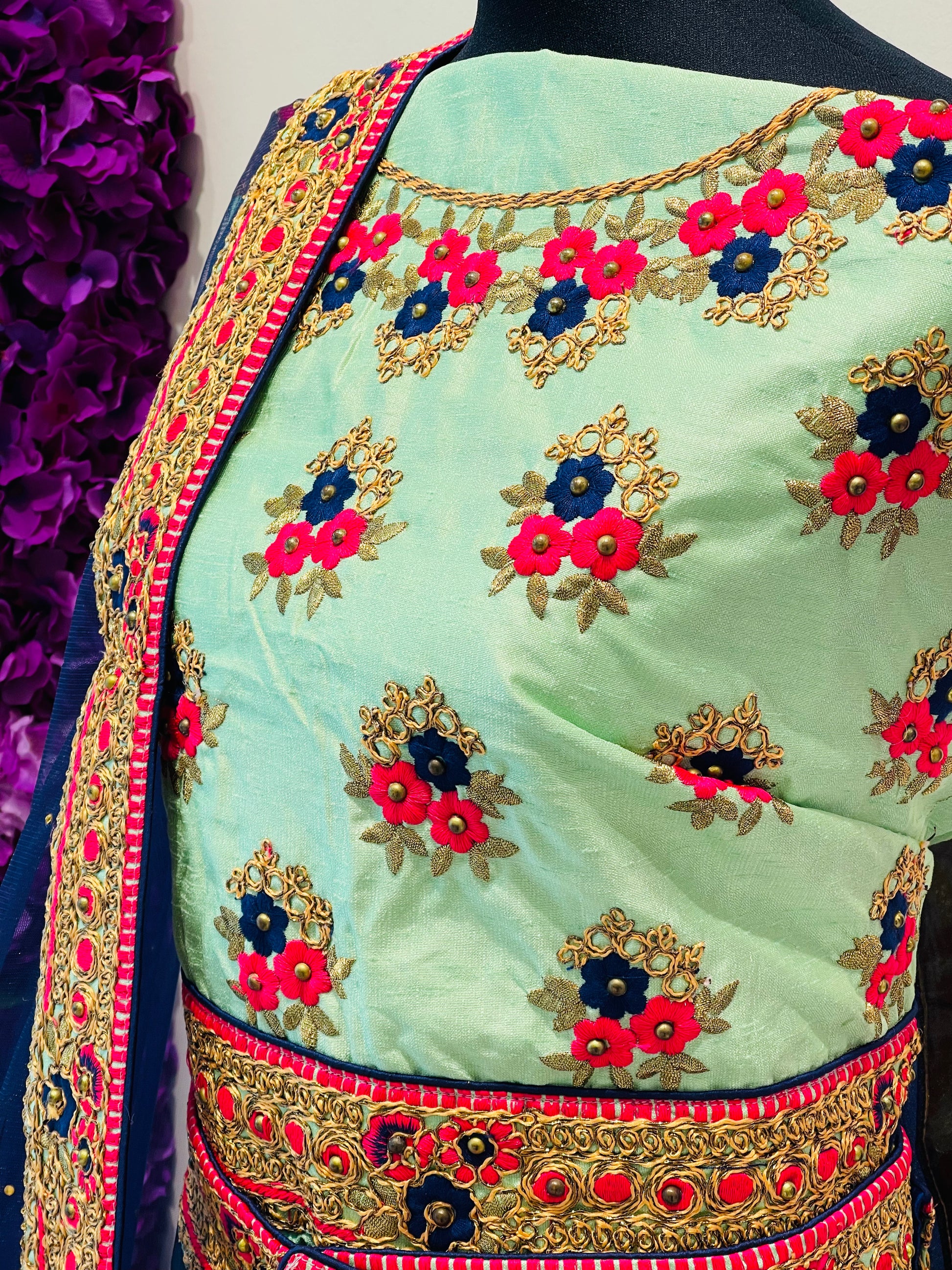 A close-up of a mint green embroidered blouse , decorated with red, navy, and gold floral motifs and a matching embroidered border.