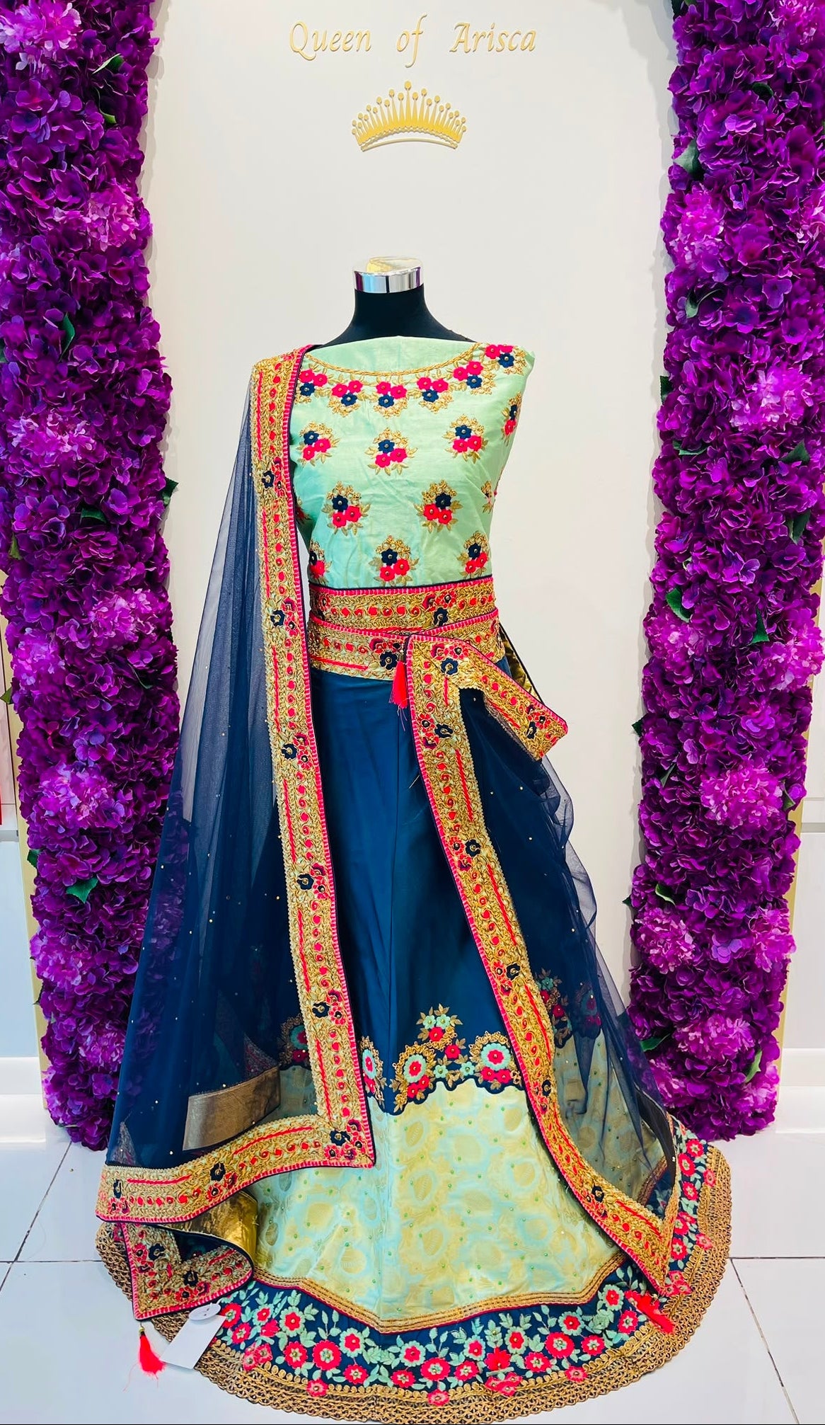 A traditional Indian lehenga dress . The outfit features a light green  with colorful floral embroidery, a navy blue skirt with gold and multicolor border details, and a sheer navy dupatta.