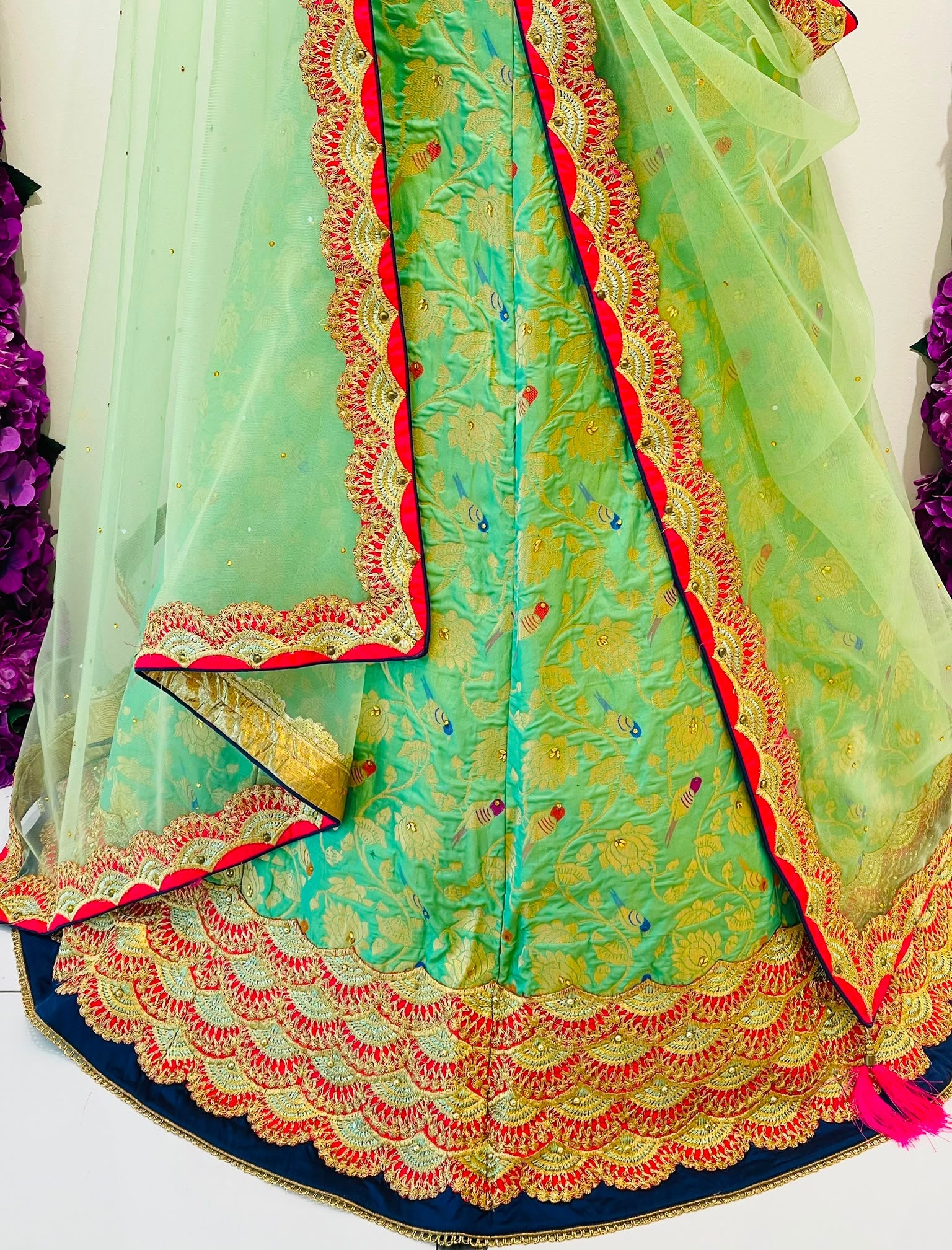 A close-up of a traditional Indian outfit featuring a mint green embroidered skirt with a navy blue scalloped hem, a red and gold embroidered border, and a matching sheer mint green dupatta draped over the side.