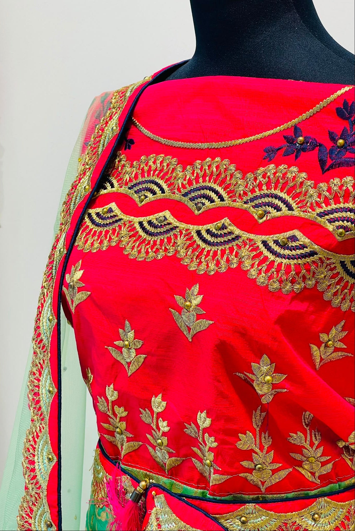 A close-up of a bright red traditional lehenga blouse, featuring ornate gold and navy floral embroidery and a sheer light green border with a gold patterned edge.