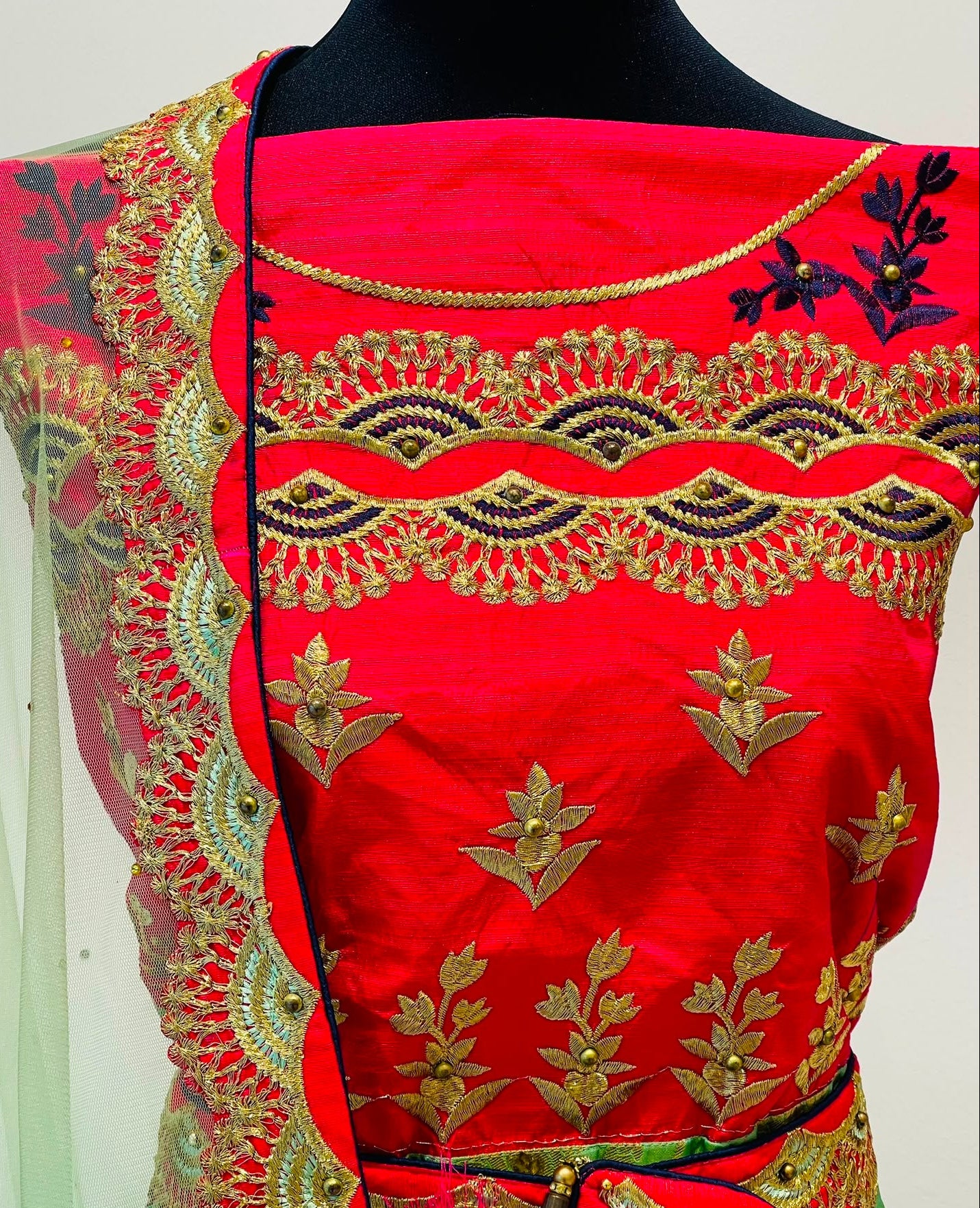 A close-up of a red traditional lehenga
featuring ornate gold and black embroidery, a matching red blouse panel, and a green dupatta with gold border detailing.