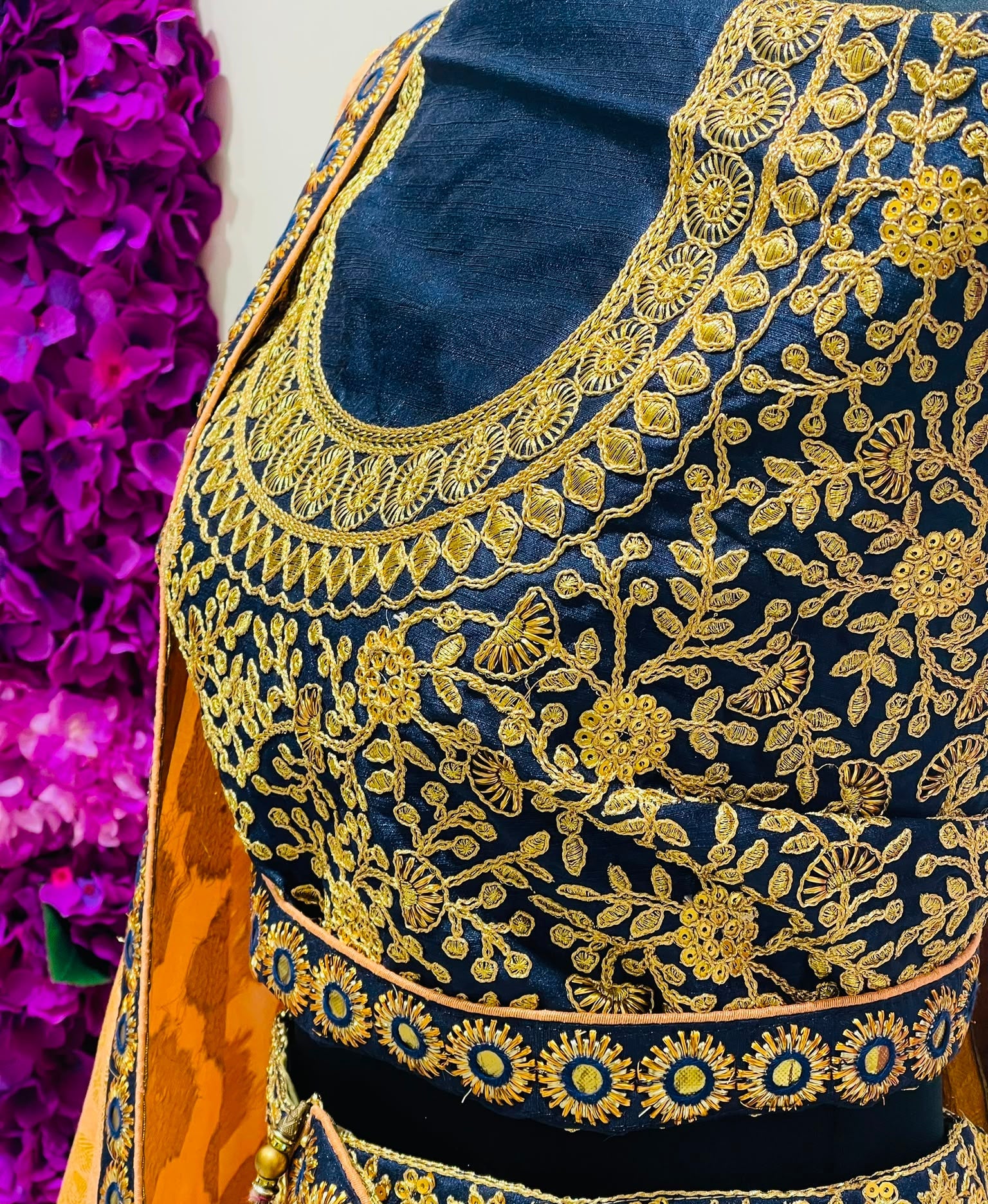 A close-up of a navy blue embroidered traditional lehenga blouse, featuring intricate gold floral embroidery and a matching orange dupatta draped over the shoulder.