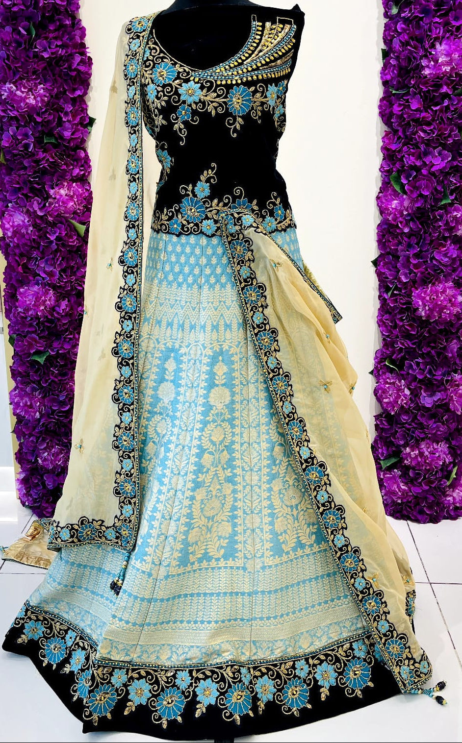 A traditional Indian lehenga featuring a navy blue sleeveless blouse with intricate gold and turquoise embroidery, a light blue printed skirt, and a beige dupatta draped over the side.