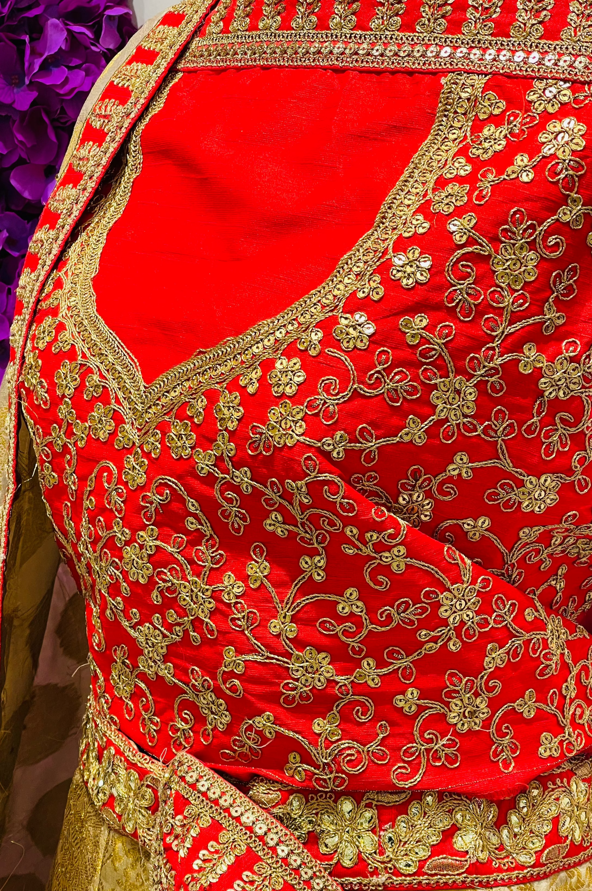 A close-up of a traditional red and gold embroidered lehenga outfit, featuring intricate gold floral embroidery on a bright red fabric, with a beige and gold patterned drape visible on the left.
