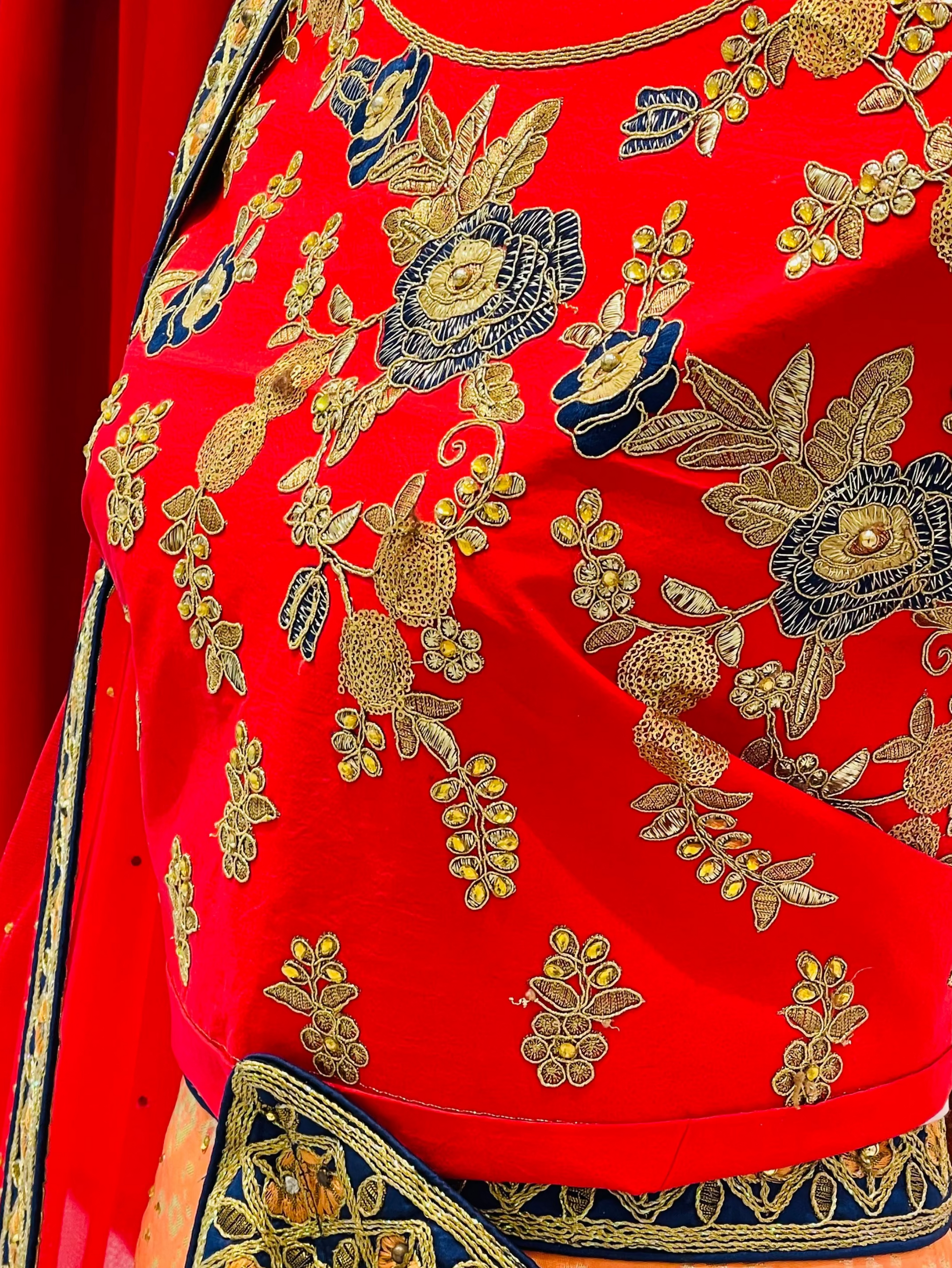 A close-up of a traditional Indian lehenga featuring a bright red  blouse with intricate gold and blue floral embroidery, paired with a red dupatta with a detailed gold border.