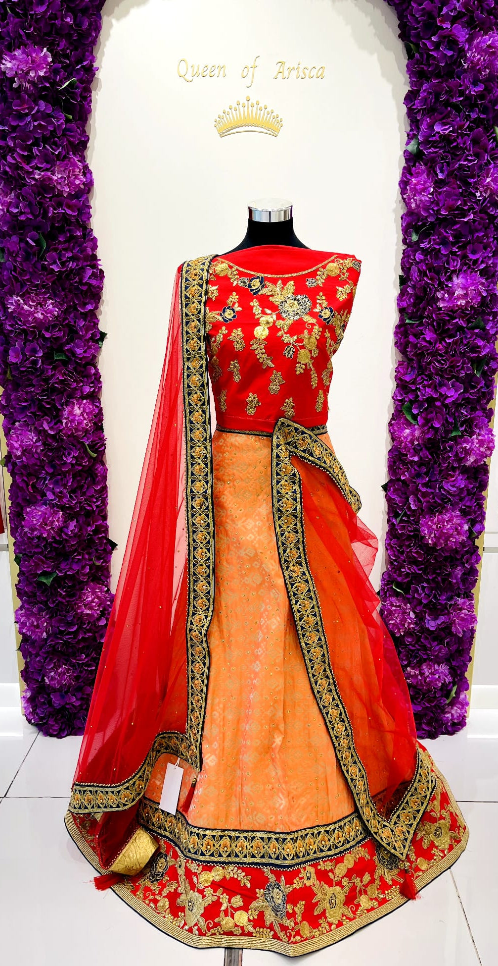 A traditional Indian lehenga . The outfit features a bright red sleeveless embroidered bodice, a peach-orange flared skirt, and a red dupatta with gold and navy blu borders. 