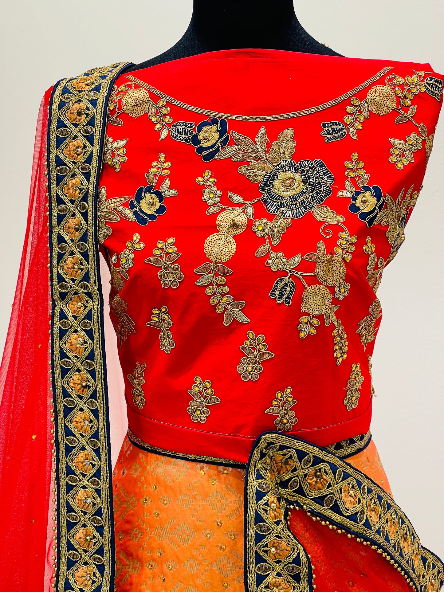 A red and orange traditional Indian lehenga .The ensemble features a bright red sleeveless blouse with intricate gold, blue, and silver floral embroidery, paired with a matching red dupatta and an orange skirt with gold detailing.