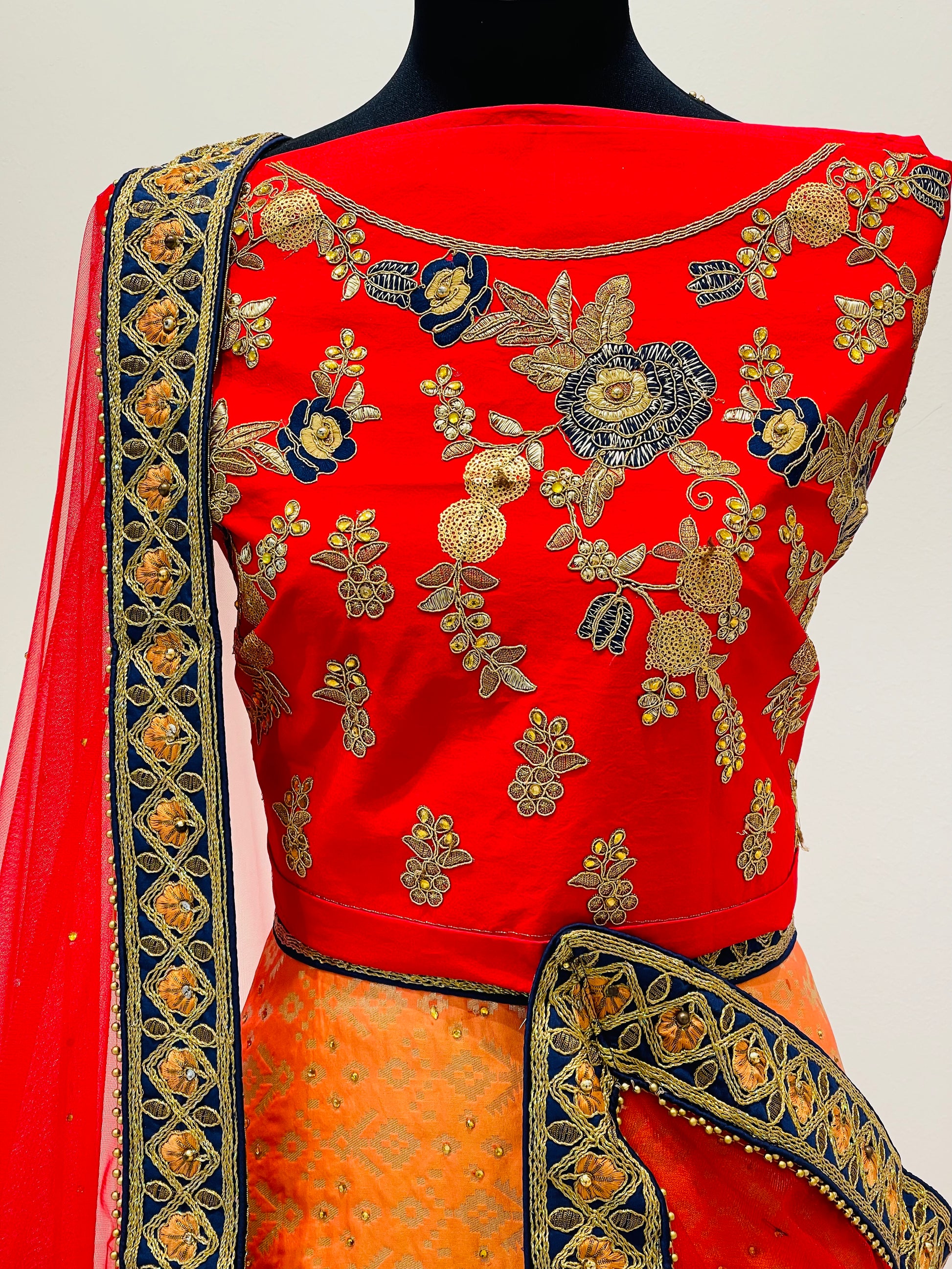 A red and orange traditional Indian lehenga .The ensemble features a bright red sleeveless blouse with intricate gold, blue, and silver floral embroidery, paired with a matching red dupatta and an orange skirt with gold detailing.