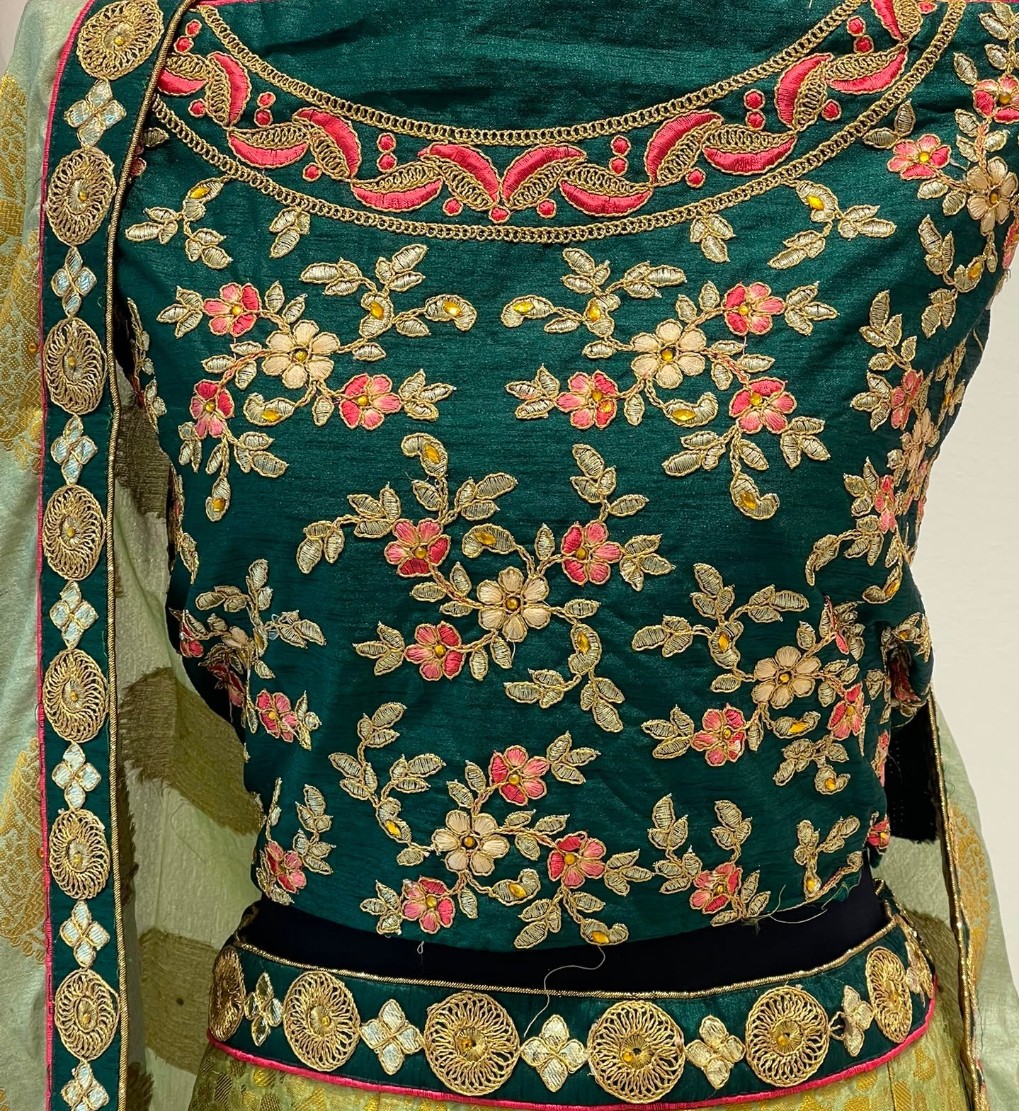 A close-up of a traditional embroidered lehenga featuring a dark teal green blouse with intricate floral embroidery in gold, pink, and white, paired with a gold patterned skirt and a matching embroidered border.