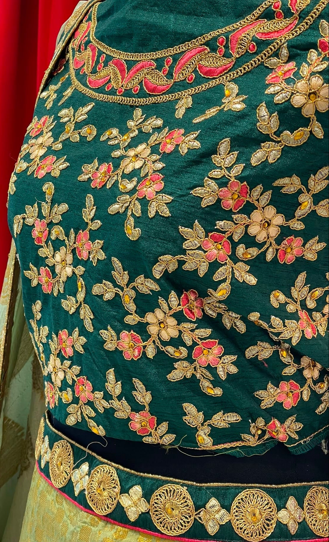 Close-up of a dark teal green embroidered blouse with floral patterns in gold, pink, and beige, paired with a gold skirt and a green dupatta with a gold border.