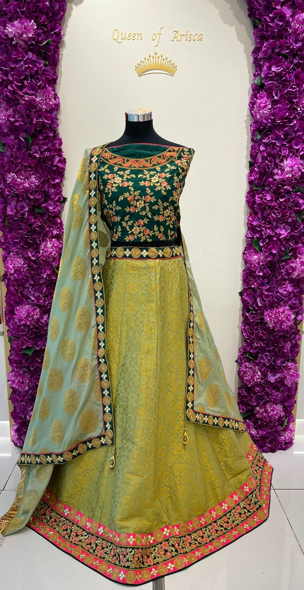 A traditional Indian lehenga choli set . The set features a dark green choli with gold floral embroidery, a light olive green skirt with a pink and gold border, and a matching light green dupatta with gold motifs.