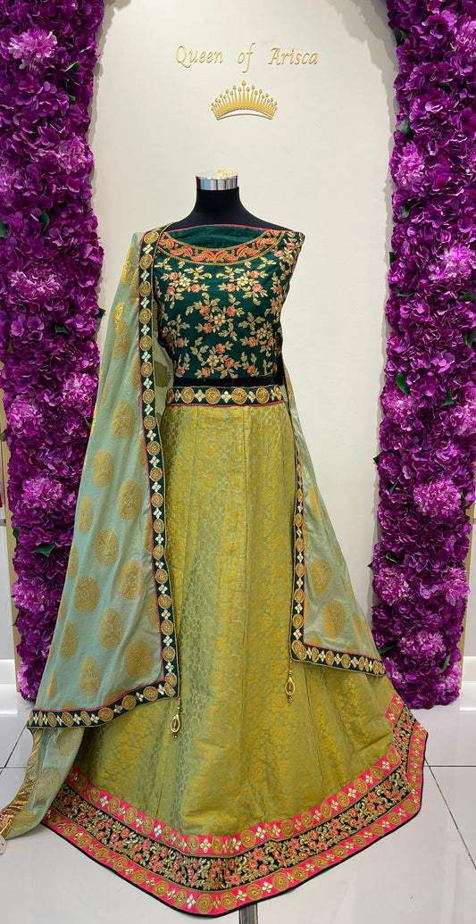 A traditional Indian lehenga choli set . The set features a dark green choli with gold floral embroidery, a light olive green skirt with a pink and gold border, and a matching light green dupatta with gold motifs.