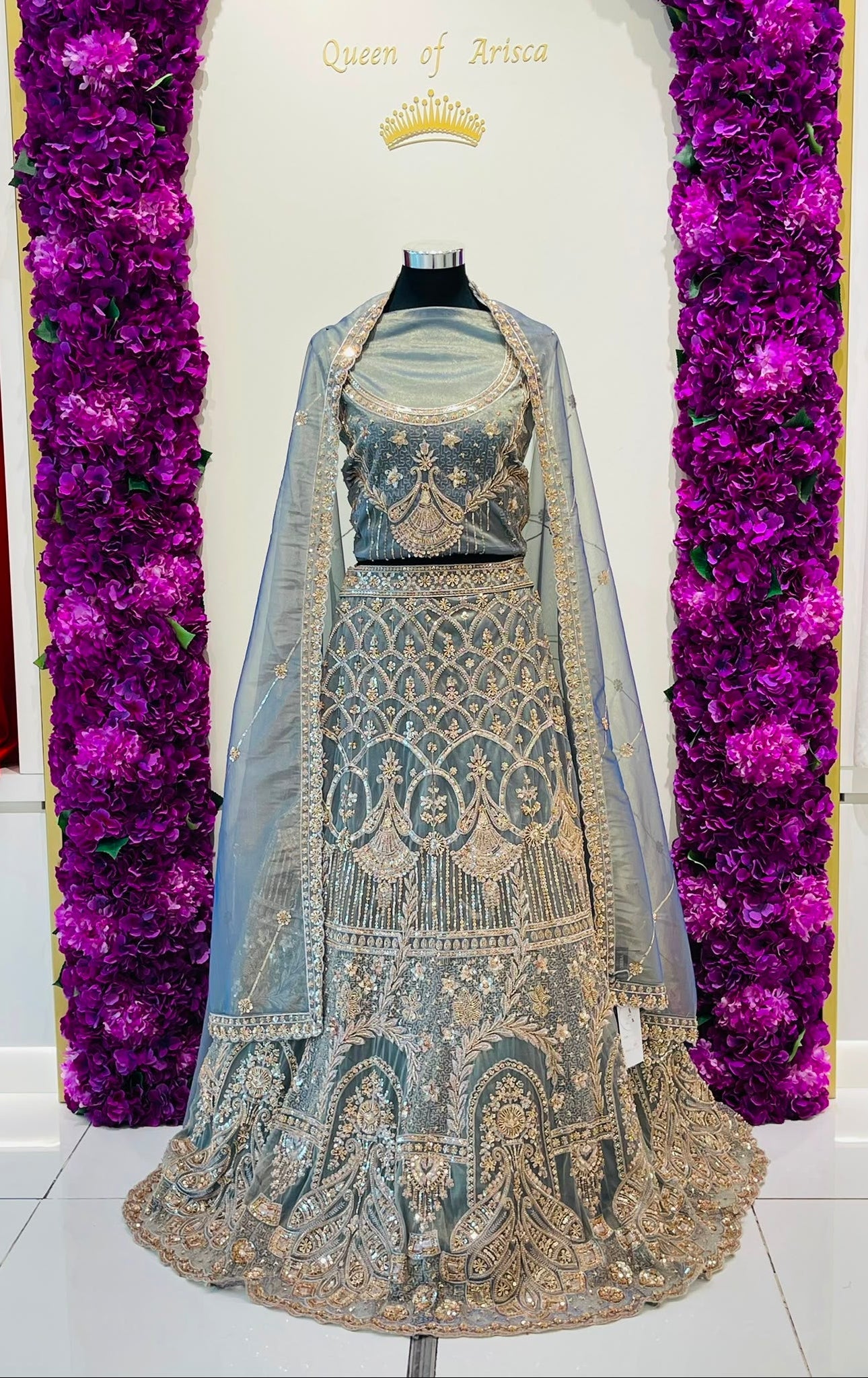 An ornate traditionl lehenga set displayed on a mannequin. The outfit features intricate gold and silver embroidery on a light blue-gray fabric, with a matching sheer dupatta draped over the shoulders.