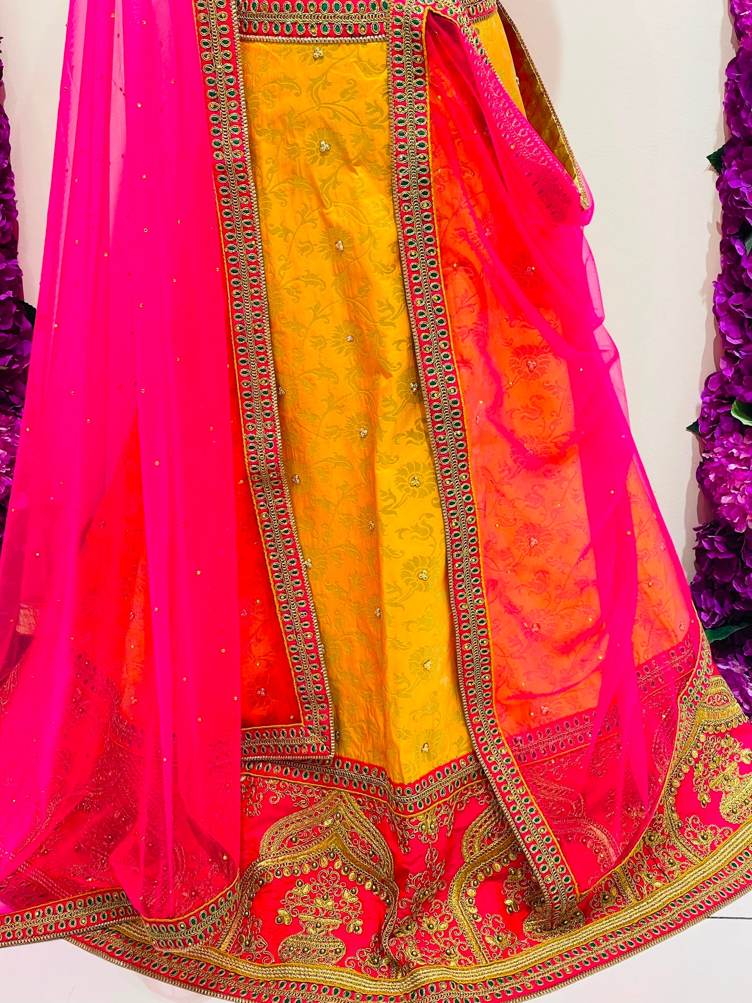A close-up of a traditional Indian outfit featuring a bright yellow embroidered kurta with a pink and orange gradient dupatta and a vibrant pink border, decorated with gold zari embroidery.