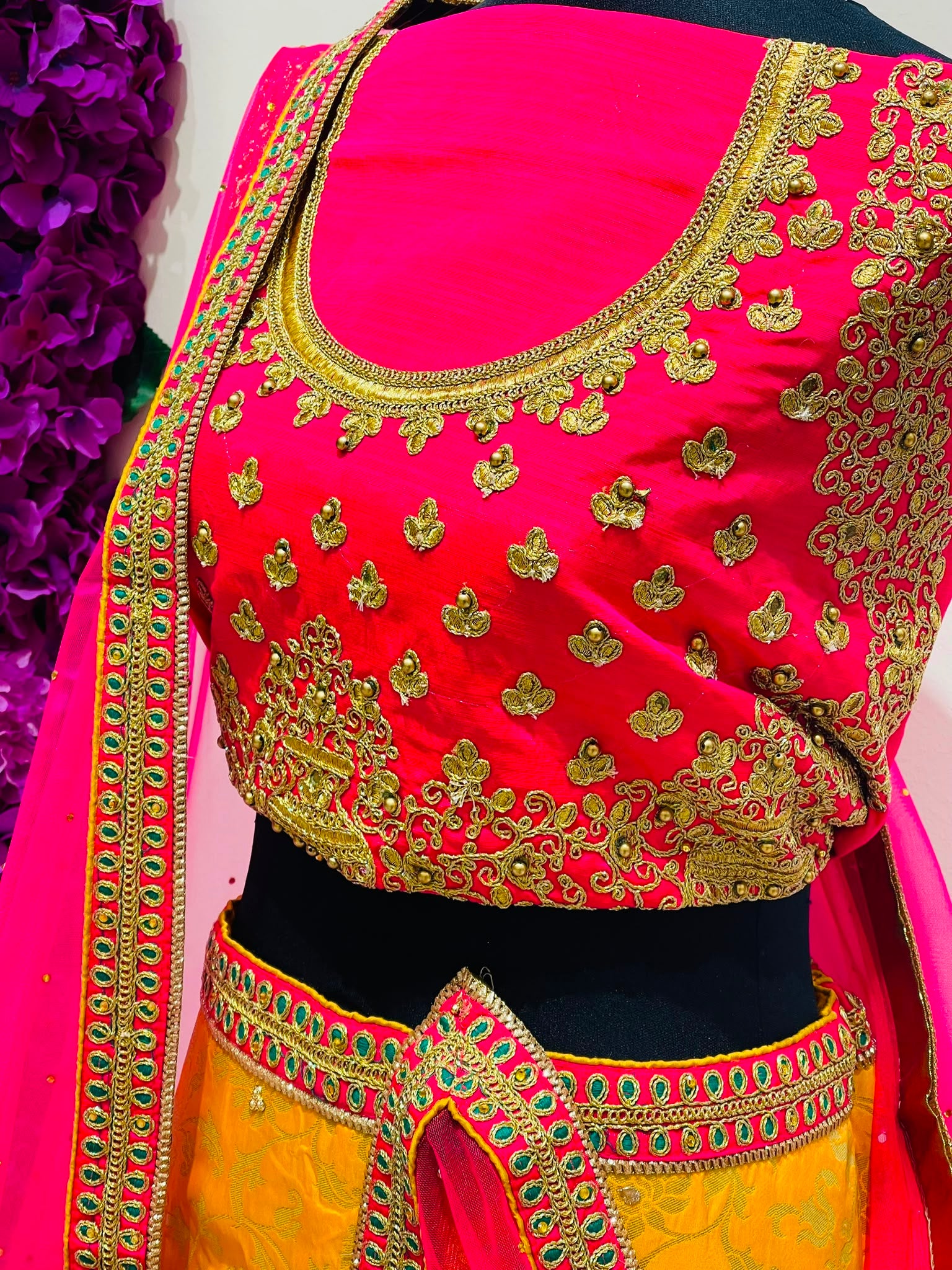 A close-up of a bright pink and yellow traditional outfit , featuring heavy gold embroidery on the blouse, a pink border with green and gold details, and a matching draped dupatta.
