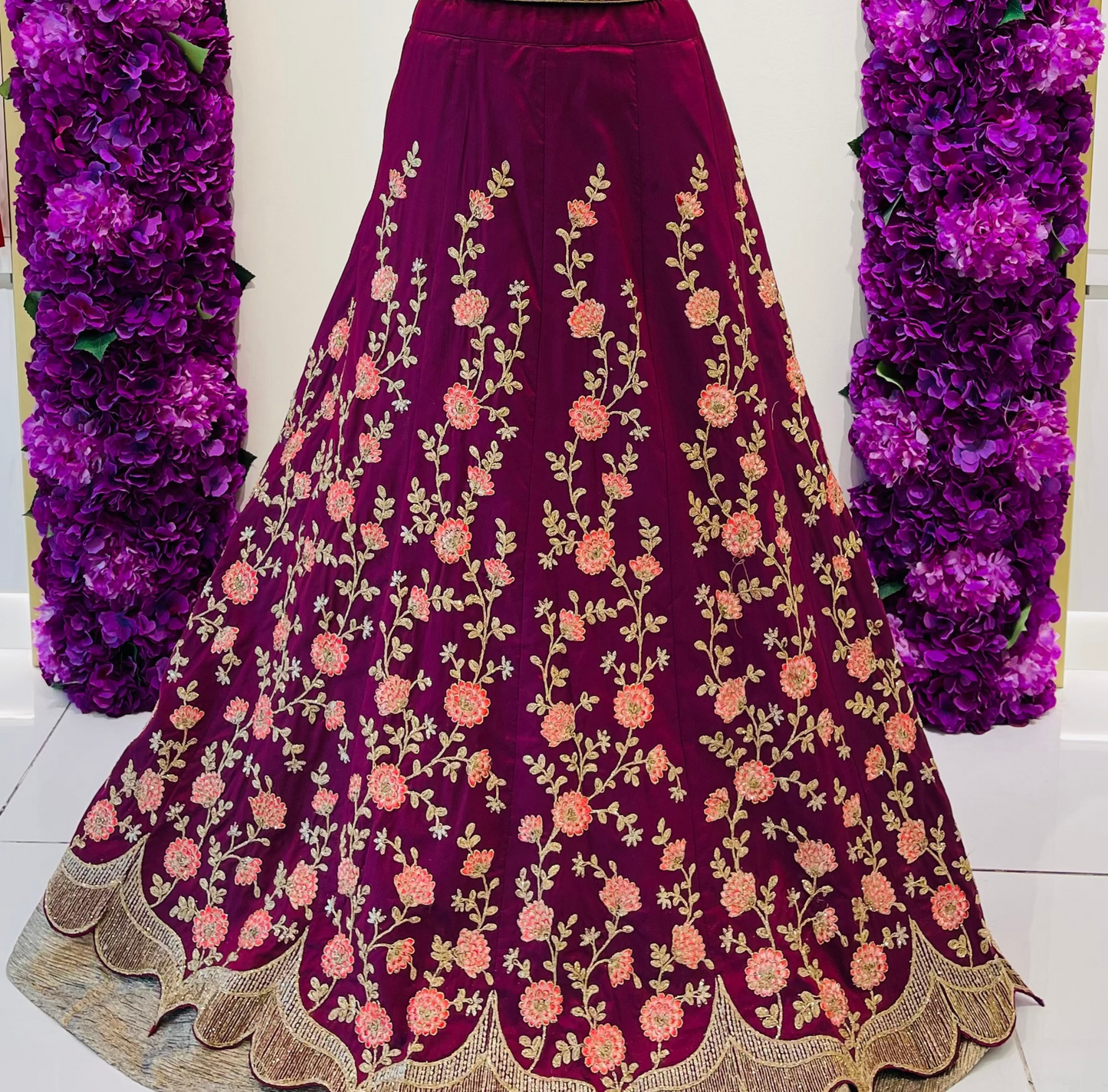 A deep magenta and purple embroidered lehenga choli skirt. The skirt features intricate gold and pink floral embroidery ,  and a scalloped gold border.