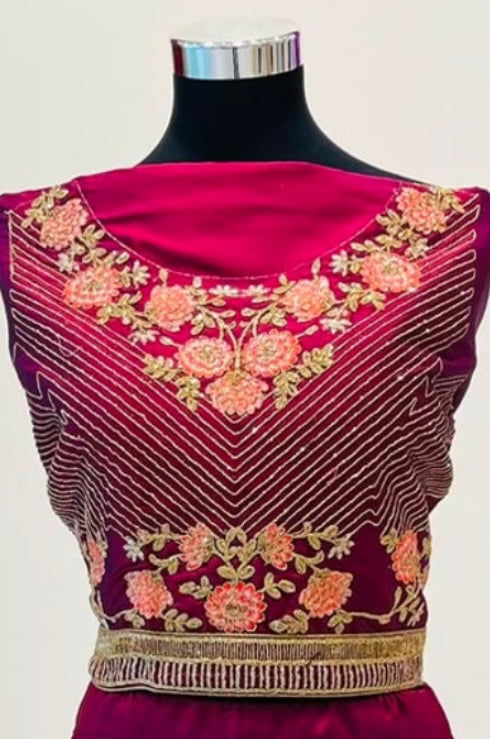 A deep magenta and purple embroidered lehenga choli set  in Arisca boutique. The dress features intricate floral embroidery in gold, pink, and beige, with a metallic gold waistband and a patterned gold border on the skirt.