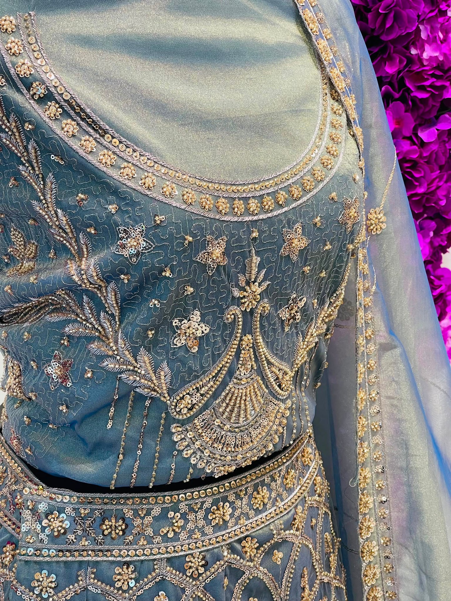 A close-up of a light blue embroidered traditional outfit with intricate gold and silver beadwork, floral patterns, and a matching sheer dupatta draped over the shoulder.